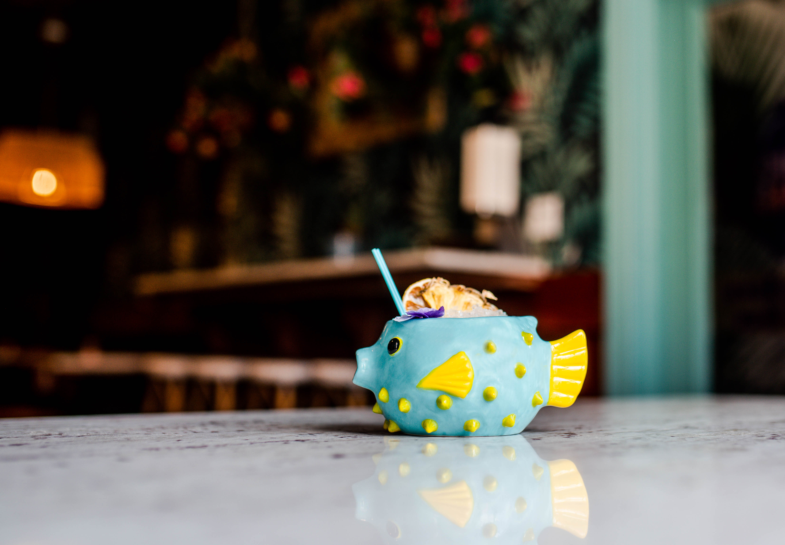 A ceramic cup shaped like a pufferfish with a blue body, yellow fins, and dots, filled with ice cream and whipped cream, placed on a white marble surface.