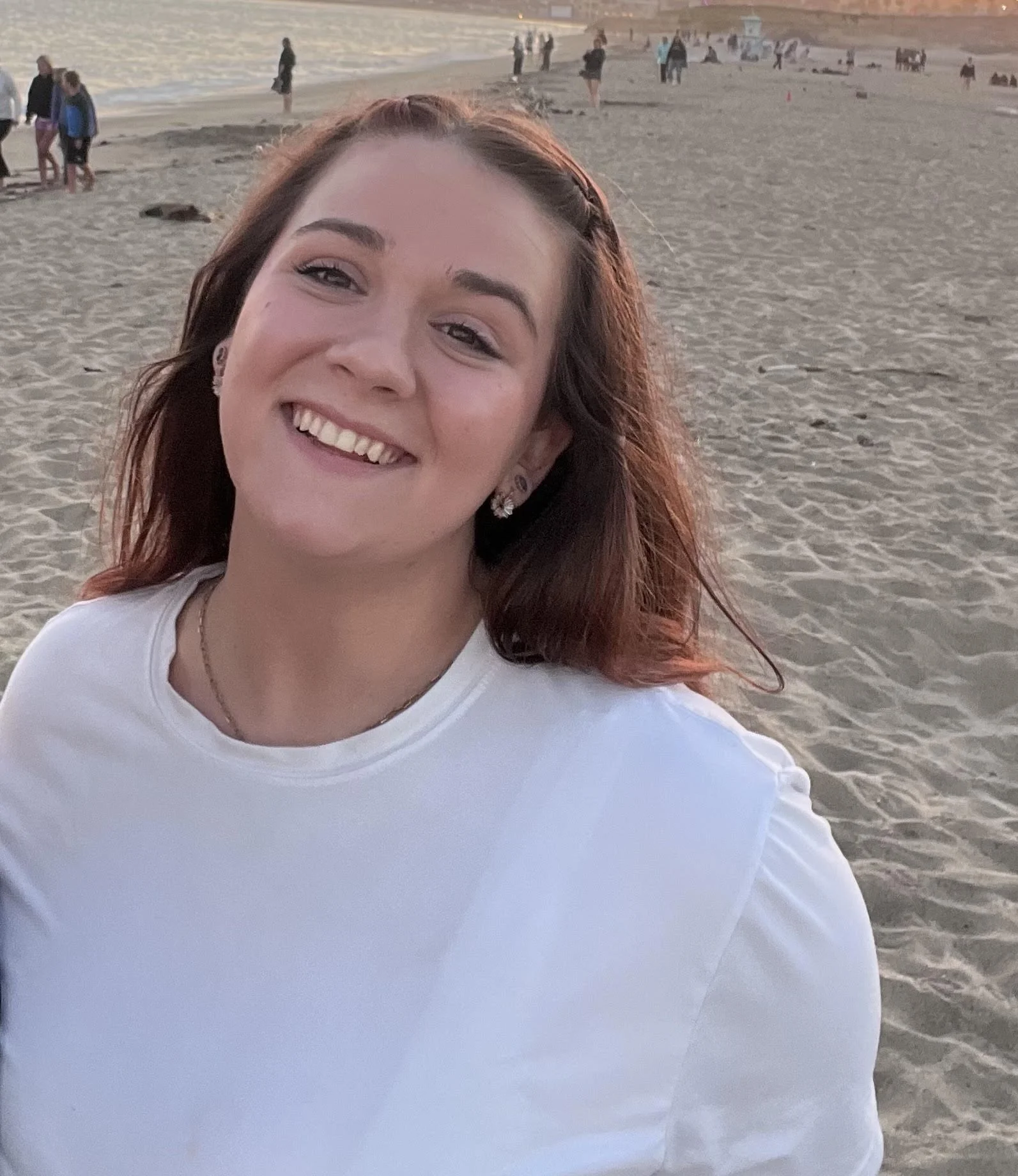 A smiling woman with red hair is at the beach, with people in the background near the shoreline and a sunset over the water.