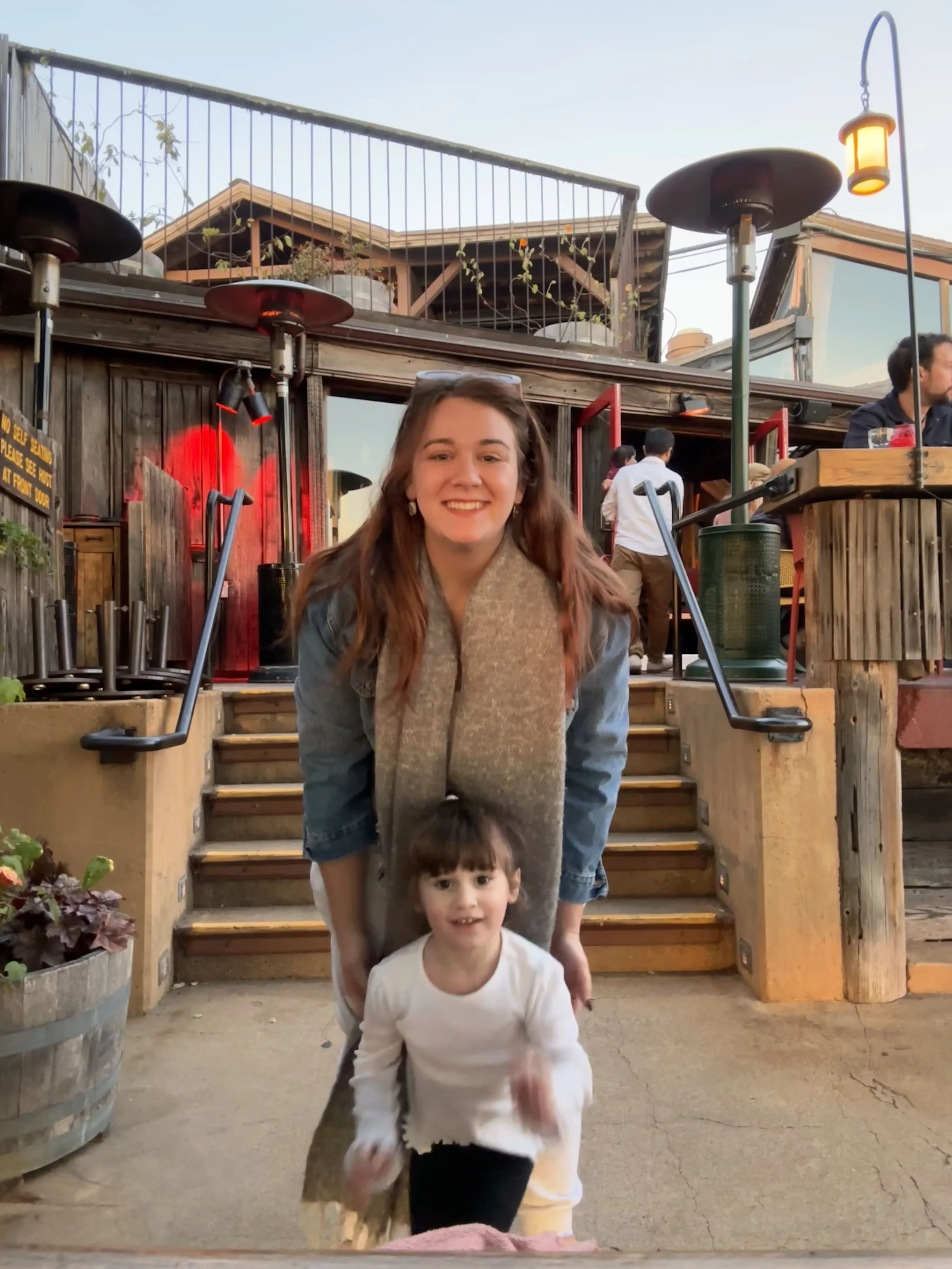 A woman with long brown hair and a gray scarf is smiling as she stands behind a young girl with dark hair and a white shirt. They are outdoors at a restaurant or bar with wooden stairs, plants, and patio heaters. Other people are visible in the background.