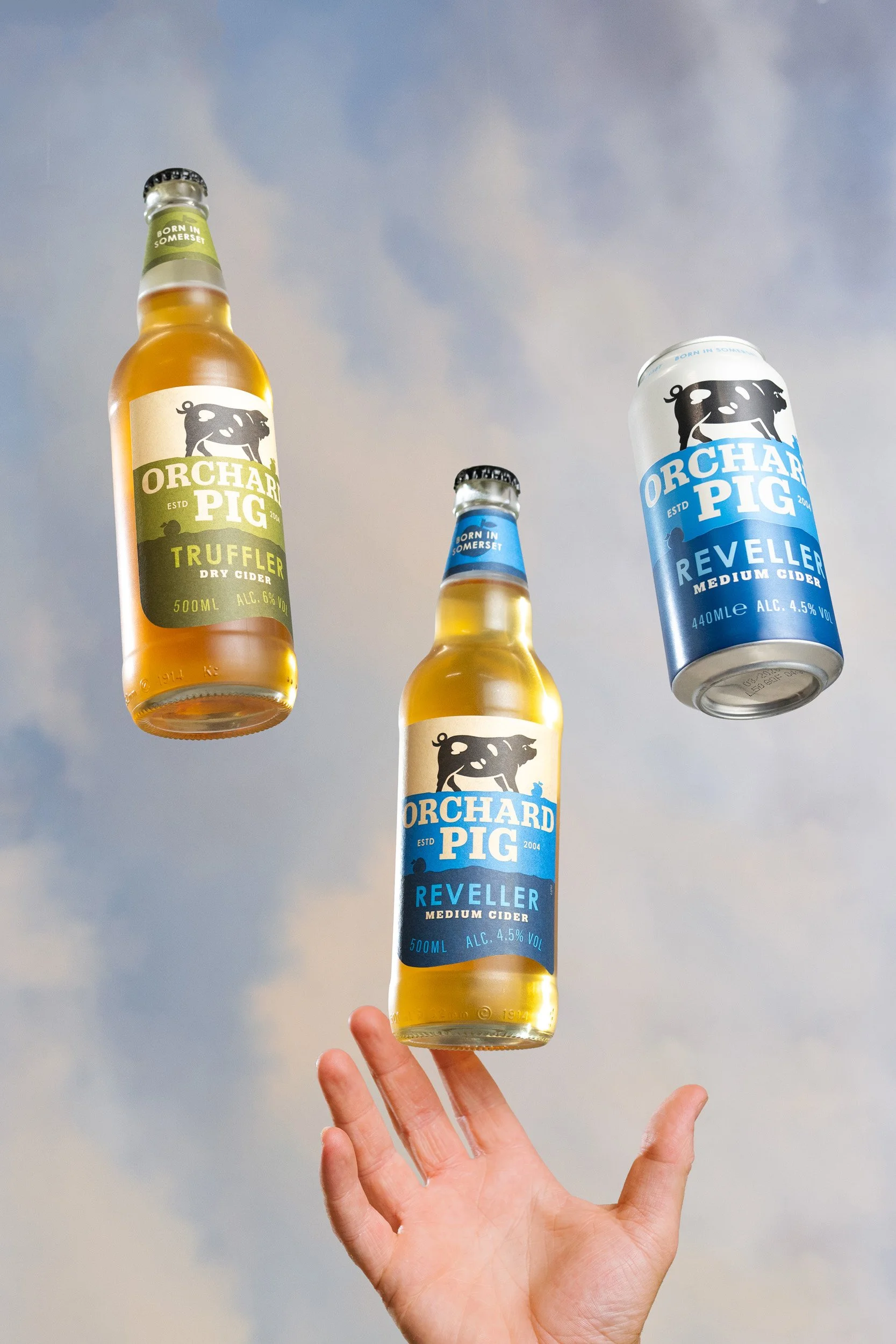 A person's hand holding three bottles of Orchard Pig cider against a cloudy sky. The bottles are labeled Truffler dry cider and Reveller medium cider.