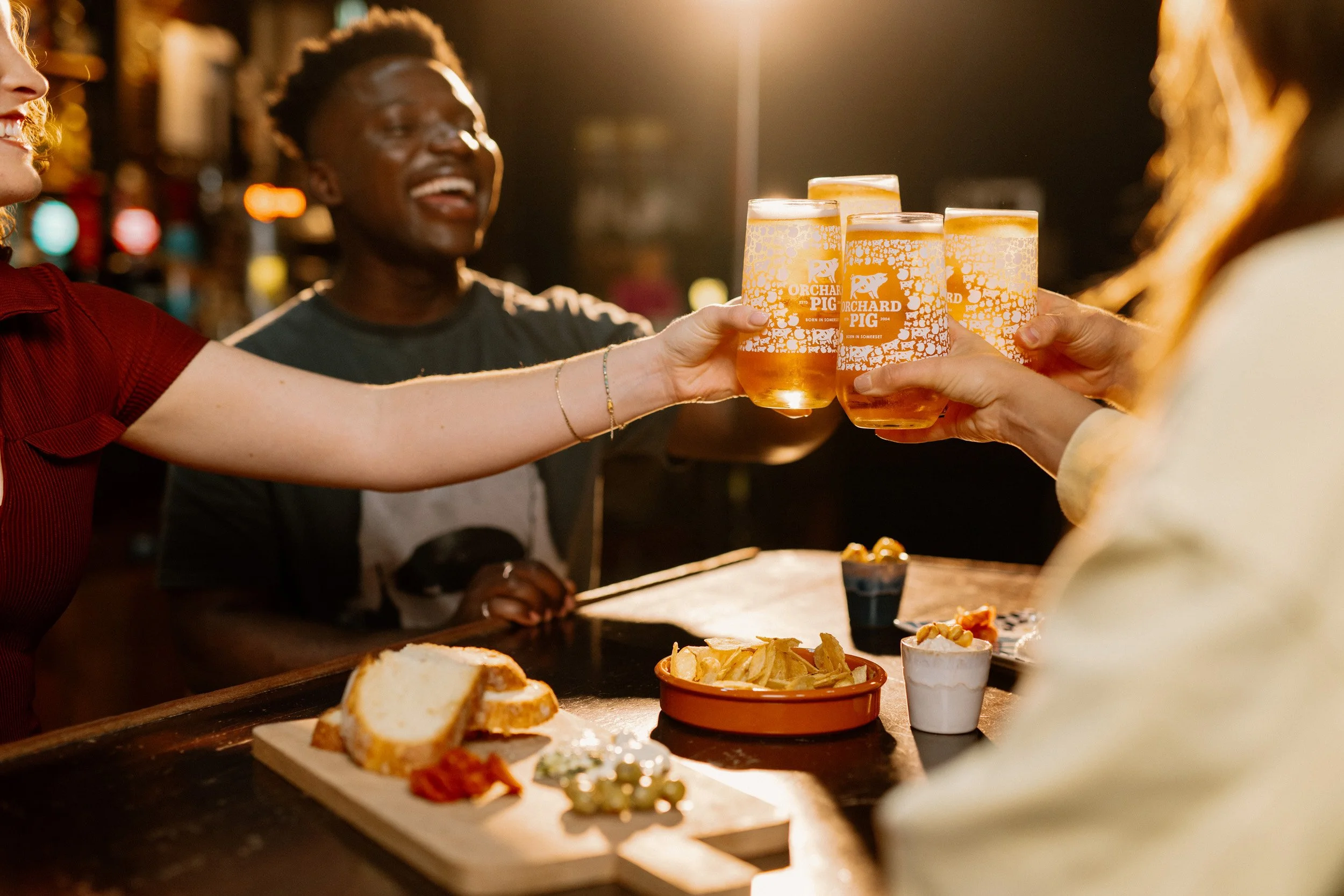 Group of friends enjoying Orchard Pig and snacks at a bar, with a focus on four glasses of beer being cheersed, and food on the table.
