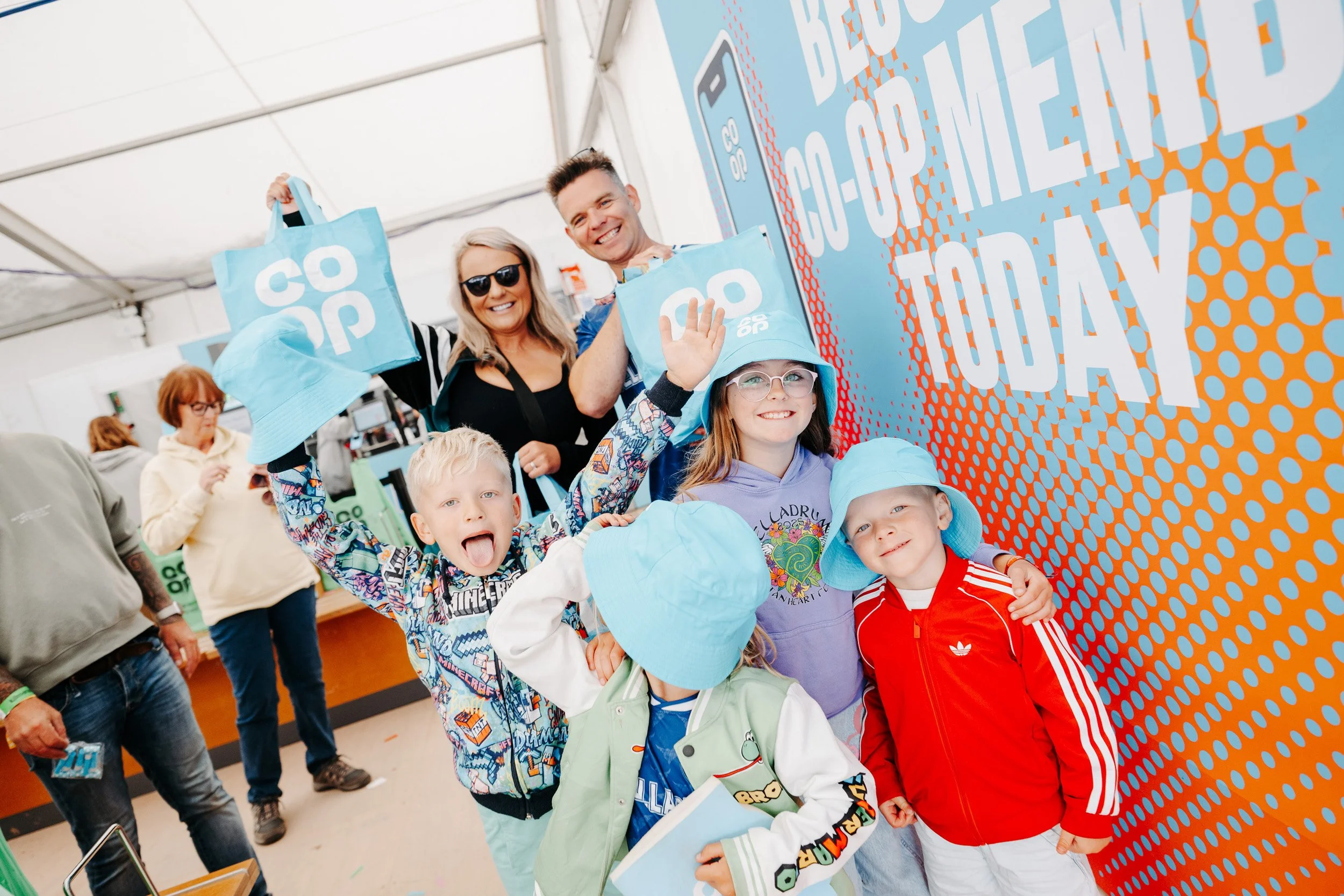 Smiling children with backpacks and light blue hats at a booth promoting sustainable shopping, with adults in the background at an indoor event.