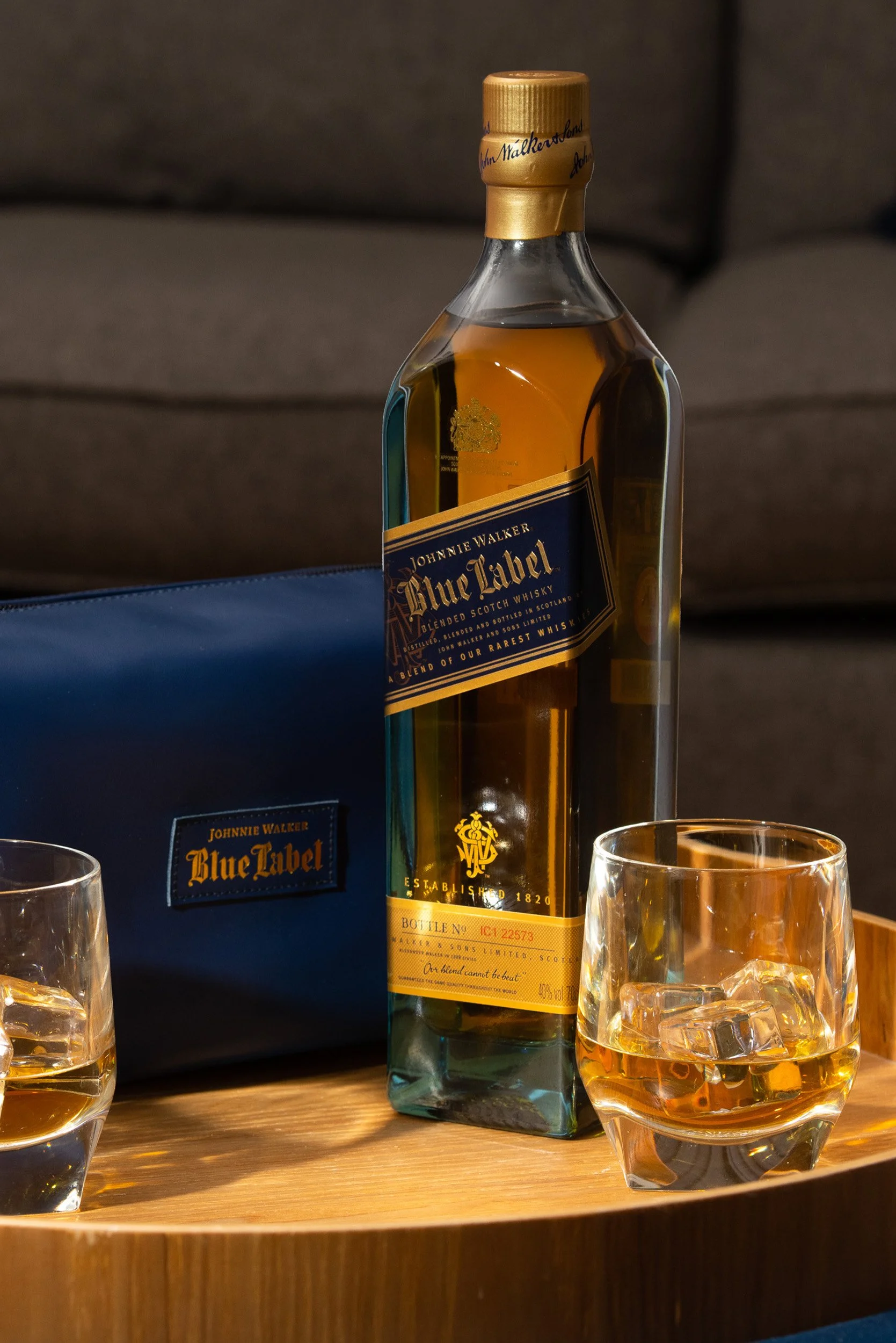 A bottle of Johnnie Walker Blue Label Scotch whisky on a wooden tray, with two glasses filled with whisky and ice in the foreground, and a dark background.