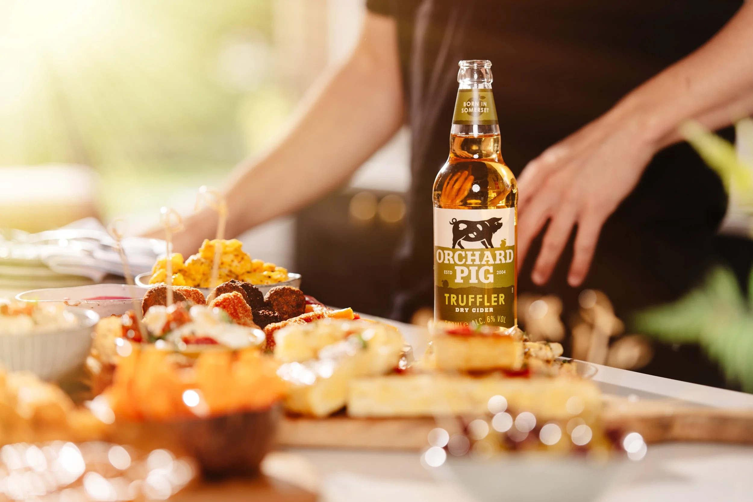 A bottle of Orchard Pig Truffler dry cider on a table surrounded by assorted appetizers and finger foods, with a person in the background.
