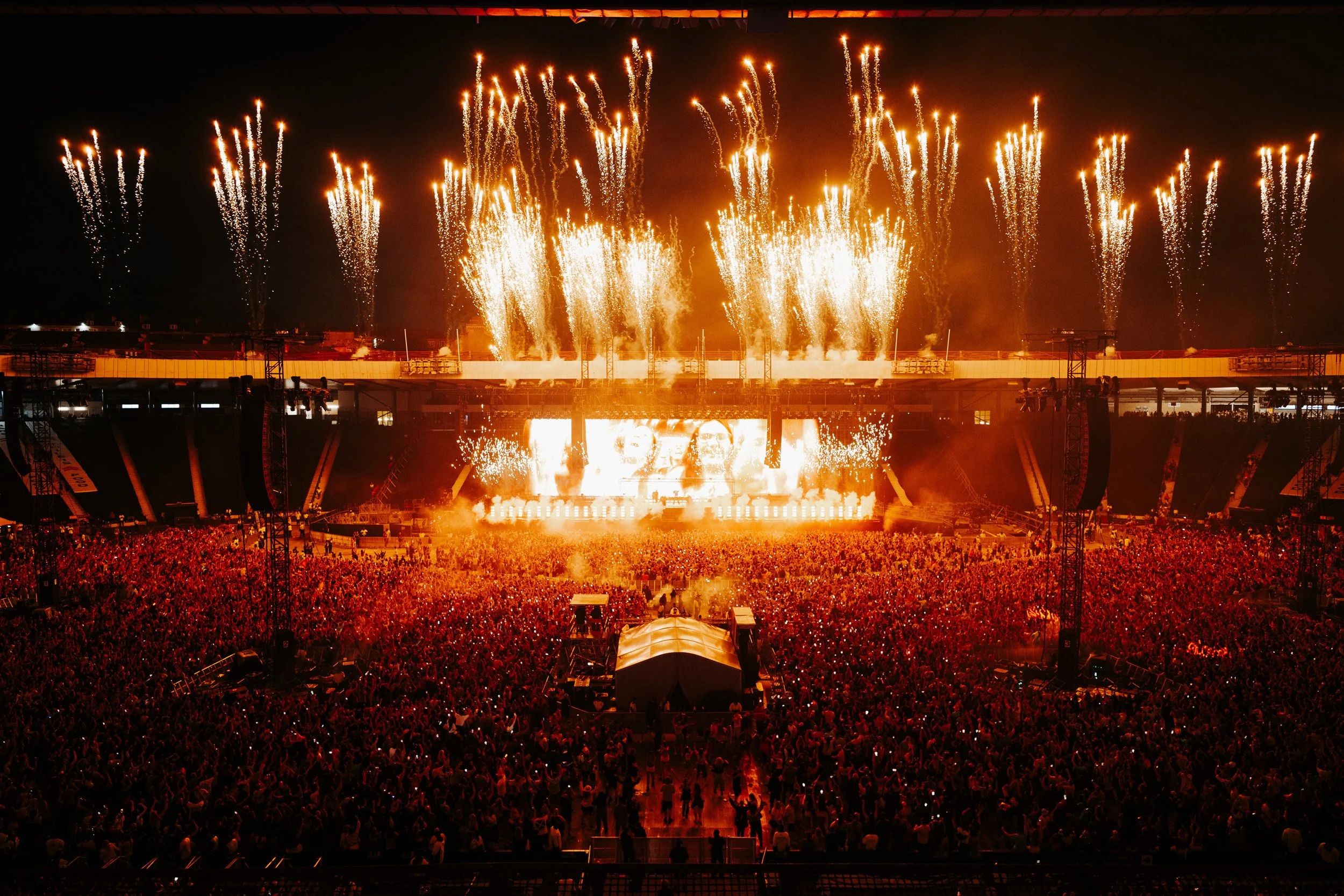 A large crowd at a concert or festival with fireworks lighting up the night sky.