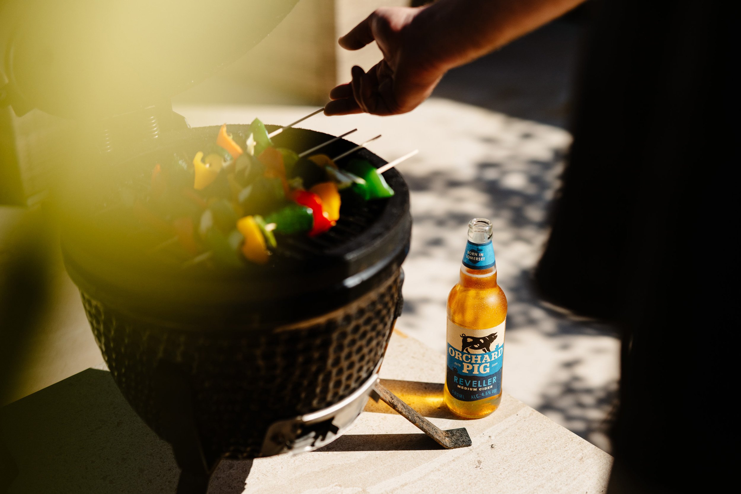 A person using a metal skewer to cook colorful bell peppers on a portable barbecue grill outdoors. A bottle of cider is on the surface beside the grill.