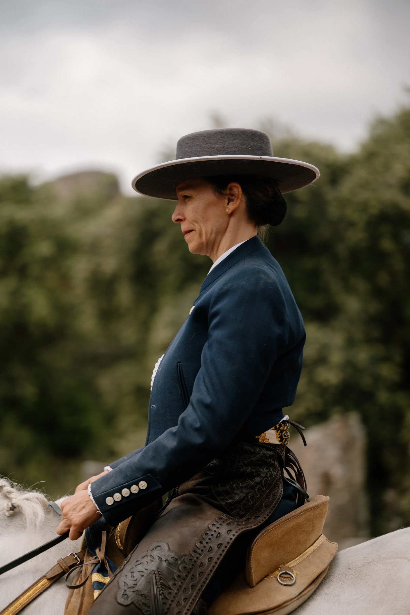 A woman dressed in traditional Mexican attire riding a horse, wearing a wide-brimmed hat and dark clothing with a blue jacket, with a wooded background.