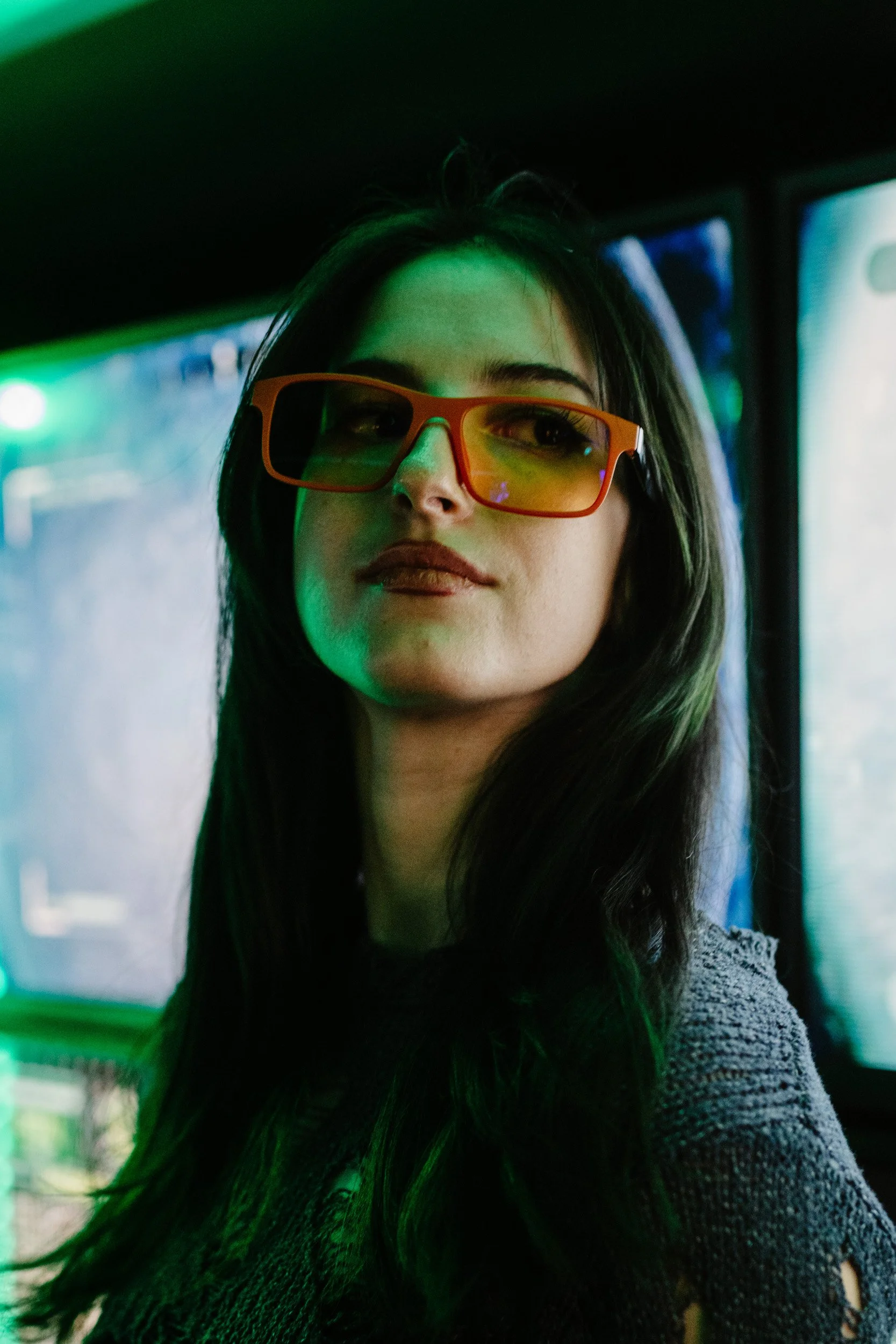 Close-up of a young woman with long dark hair wearing orange-tinted sunglasses, looking sideways, indoors with colorful lighting.