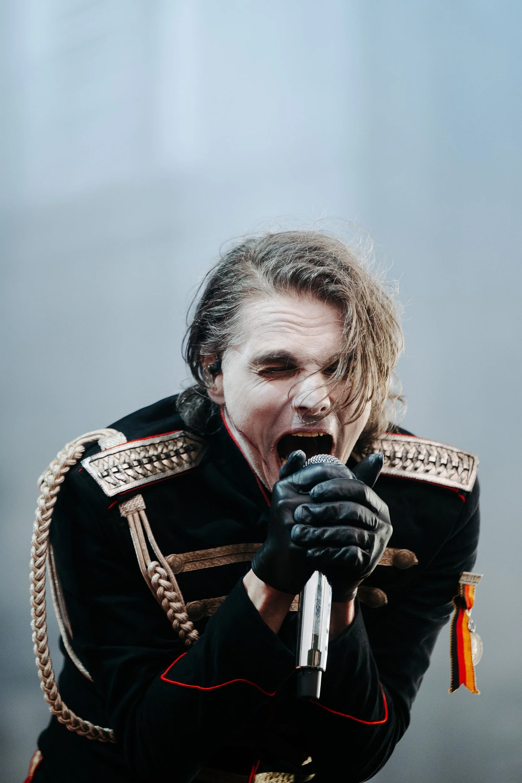 A man with long hair, dressed in a military-style uniform, passionately singing into a microphone.