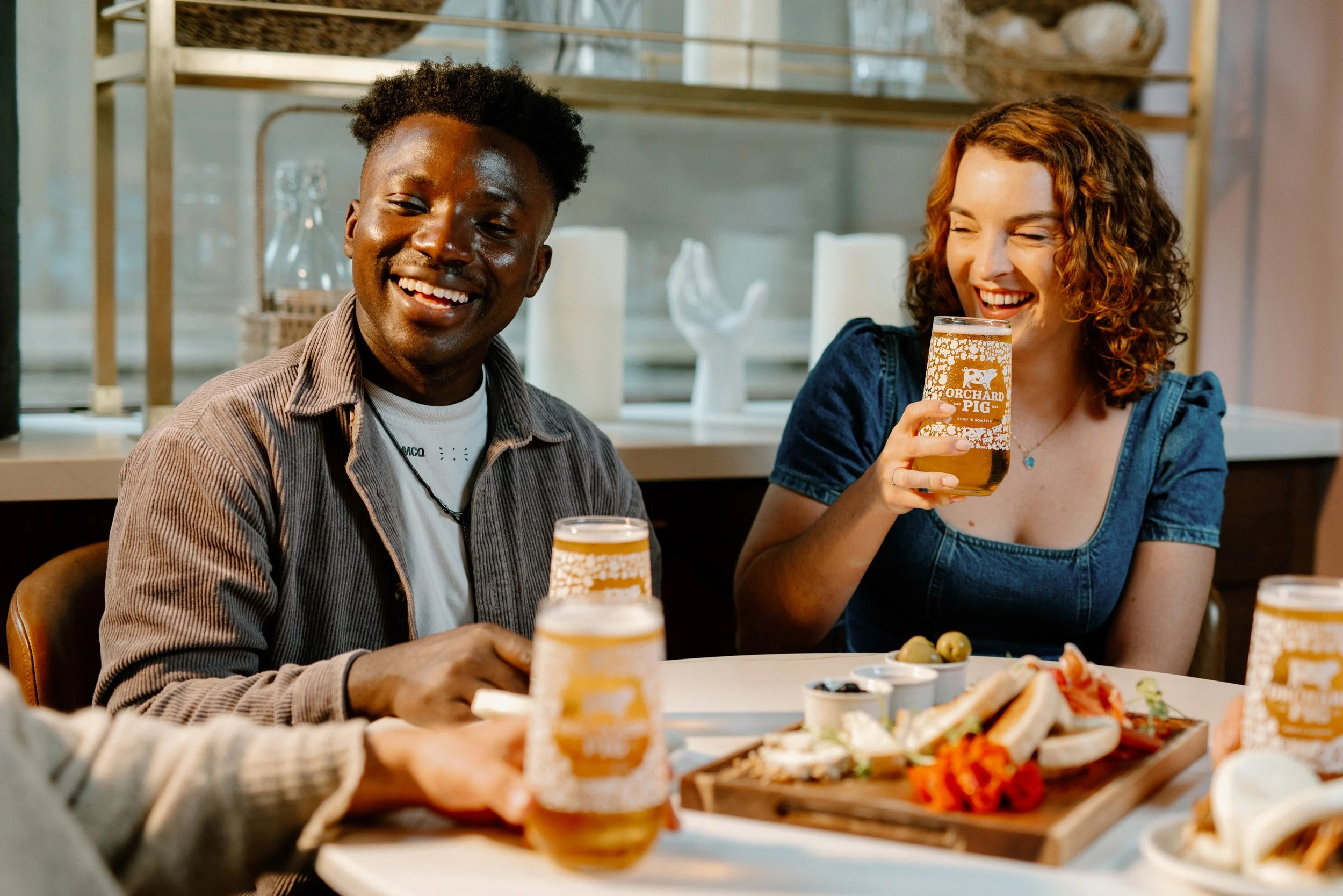 Two people smiling at a table with food and drinks, one woman holding a beer glass labeled 'Orchard Pig'.