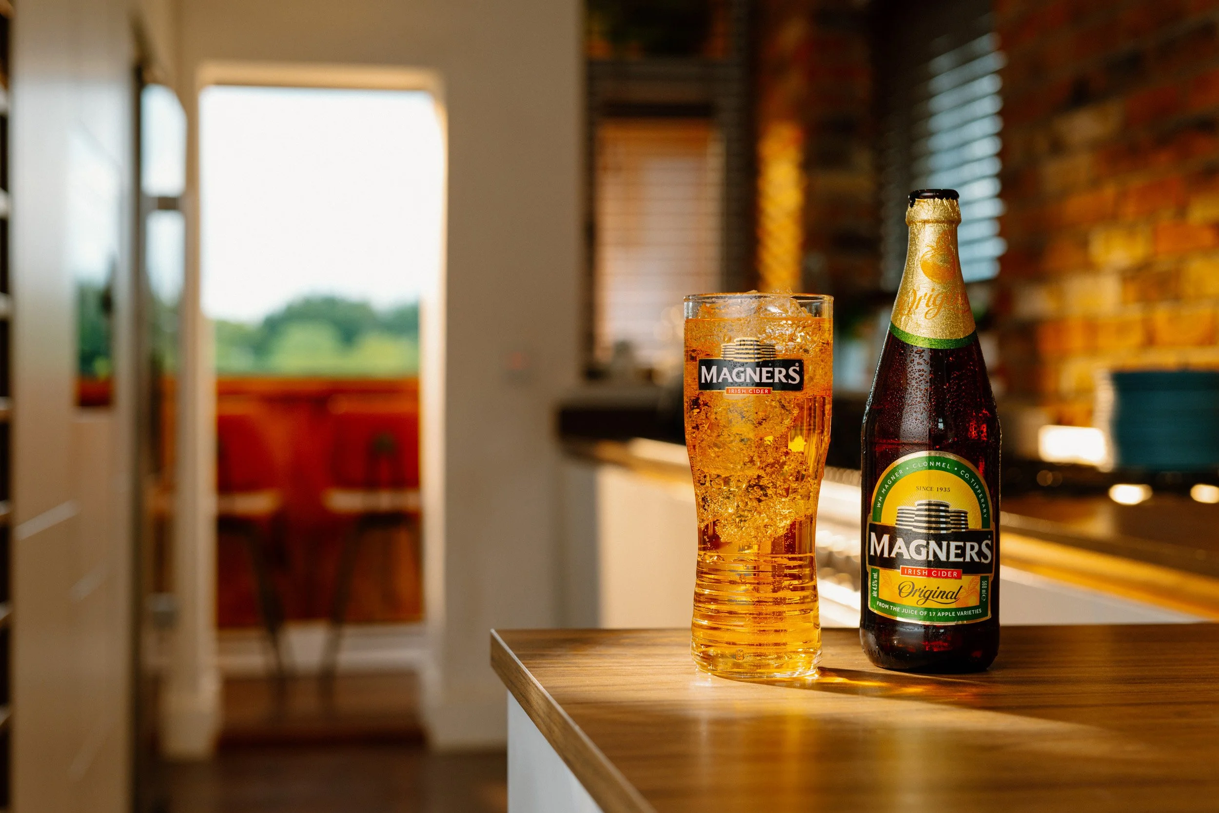 A bottle of Magners original Irish cider and a glass filled with cider on a wooden counter in a brightly lit kitchen or dining area.