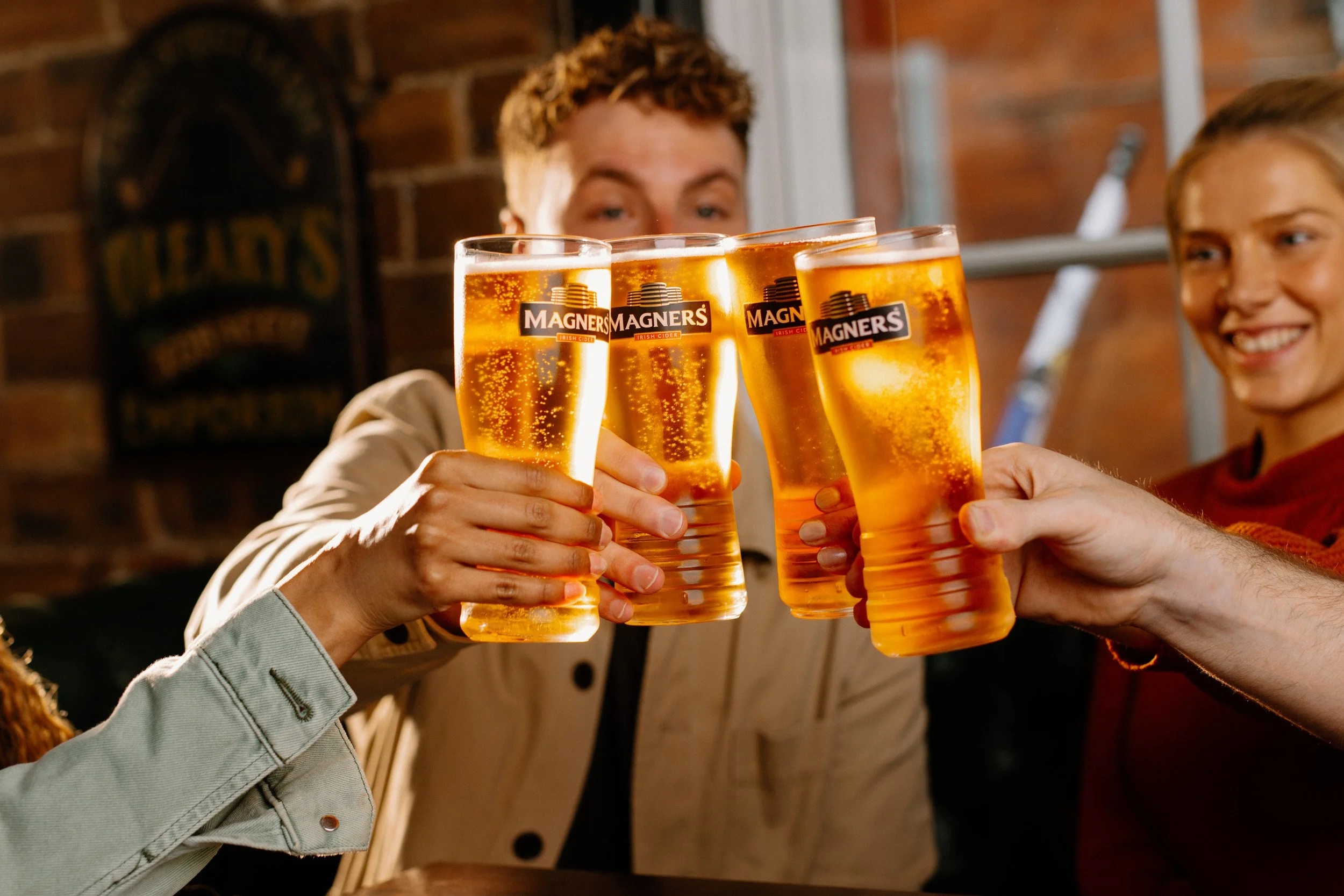 People raising glasses of amber-colored cider Magners in a toast at a bar, with a brick wall background and one of them smiling.
