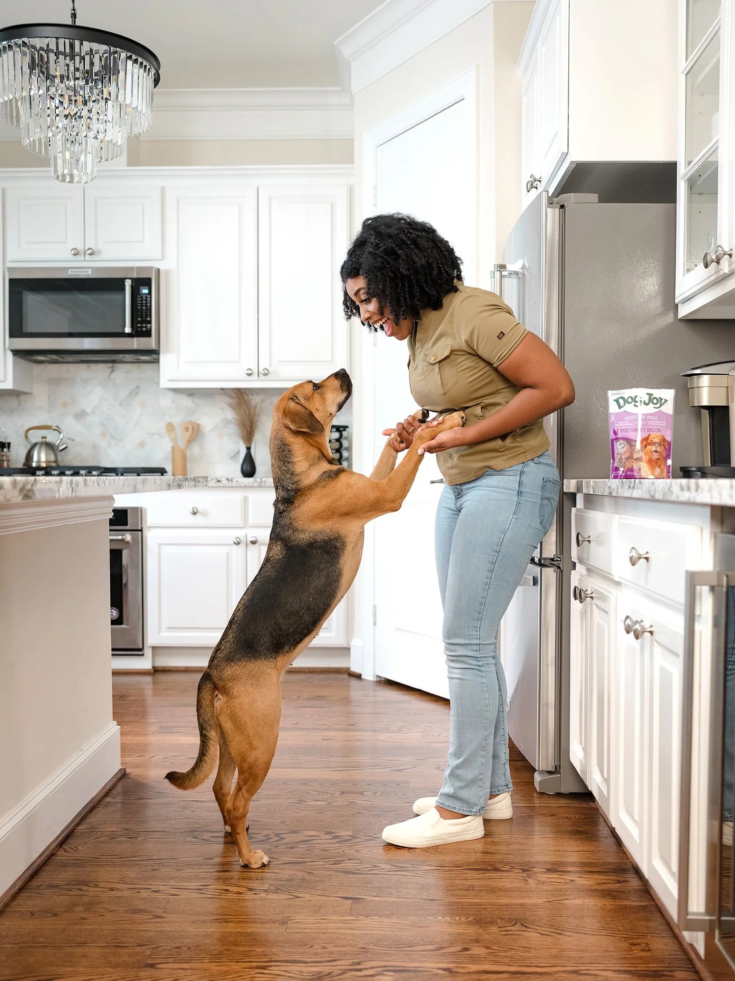 A woman and a dog playing together in a kitchen. The woman is smiling and holding the dog's paws, while the dog stands on its hind legs facing her.