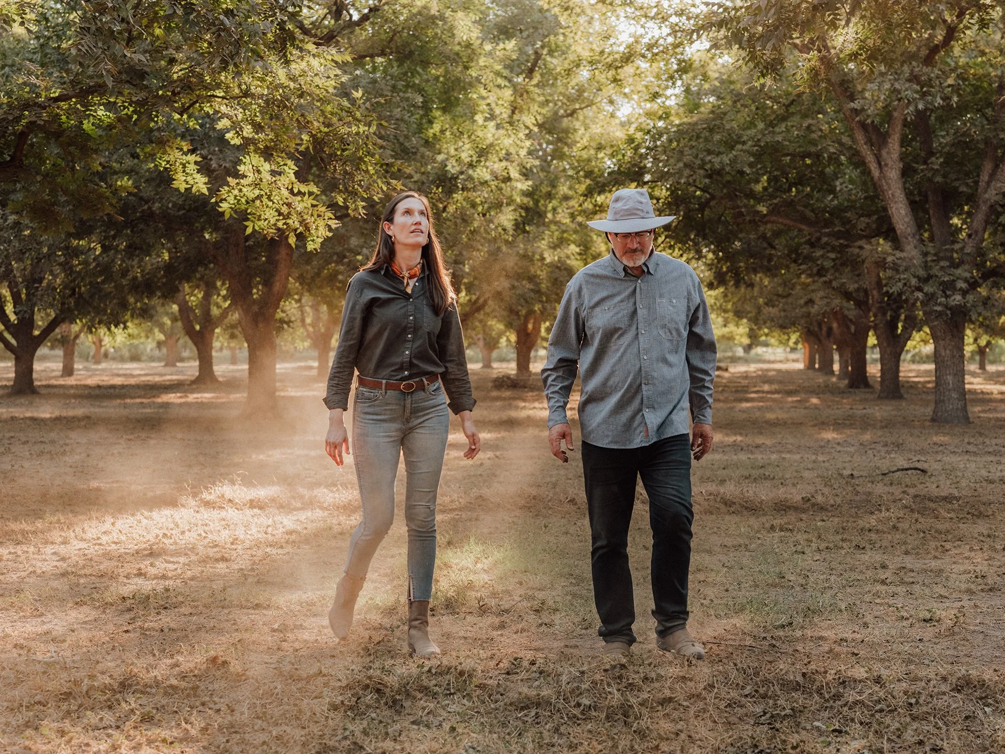 A woman and a man walking through a tree orchard during golden hour. The woman wears jeans, a dark shirt, and boots, and looks upward. The man wears a wide-brimmed hat, glasses, a gray shirt, and dark pants, and is looking downward.