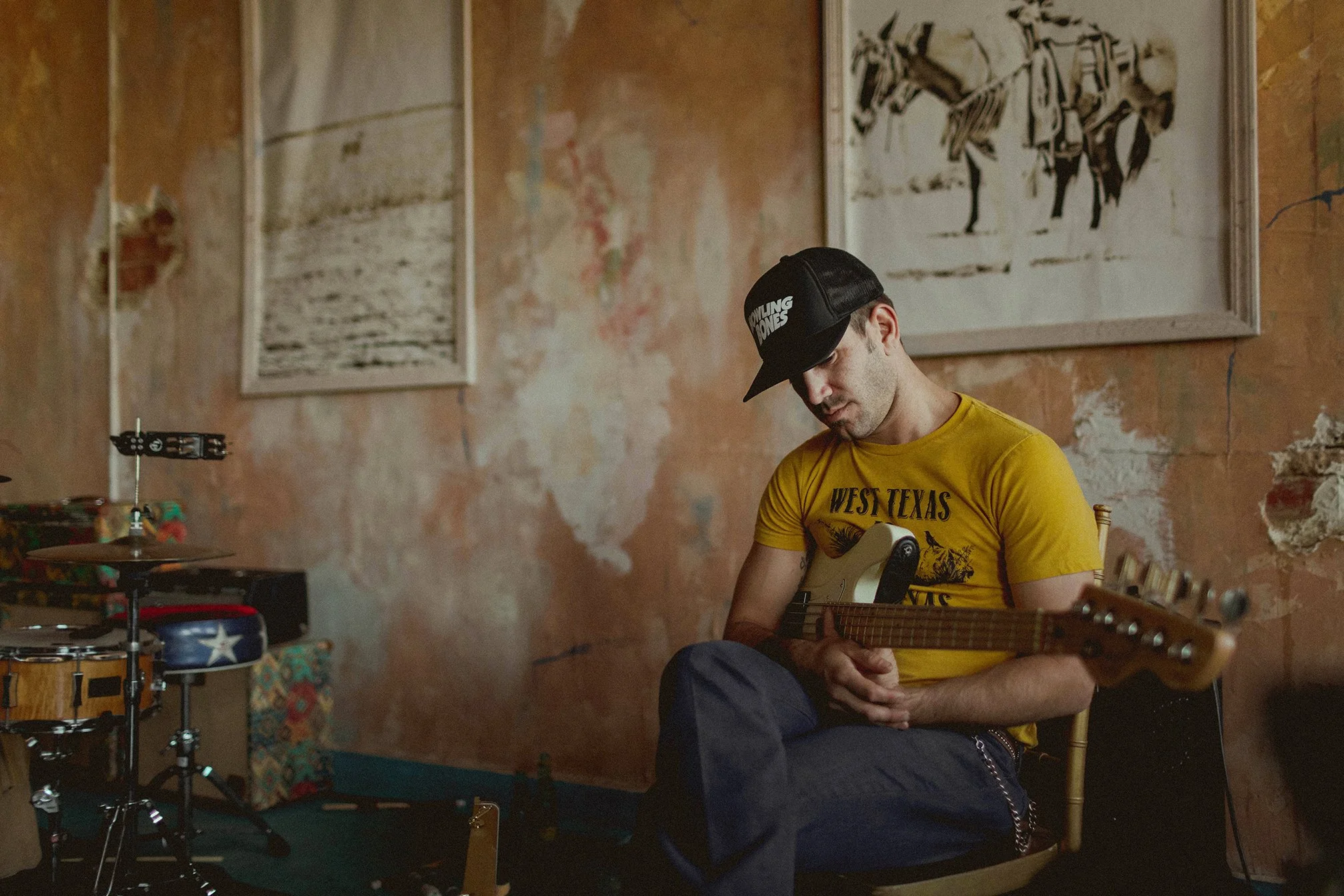 Man sitting on chair in rustic room playing electric guitar, wearing a yellow T-shirt and black baseball cap, with artwork on distressed wall in background.