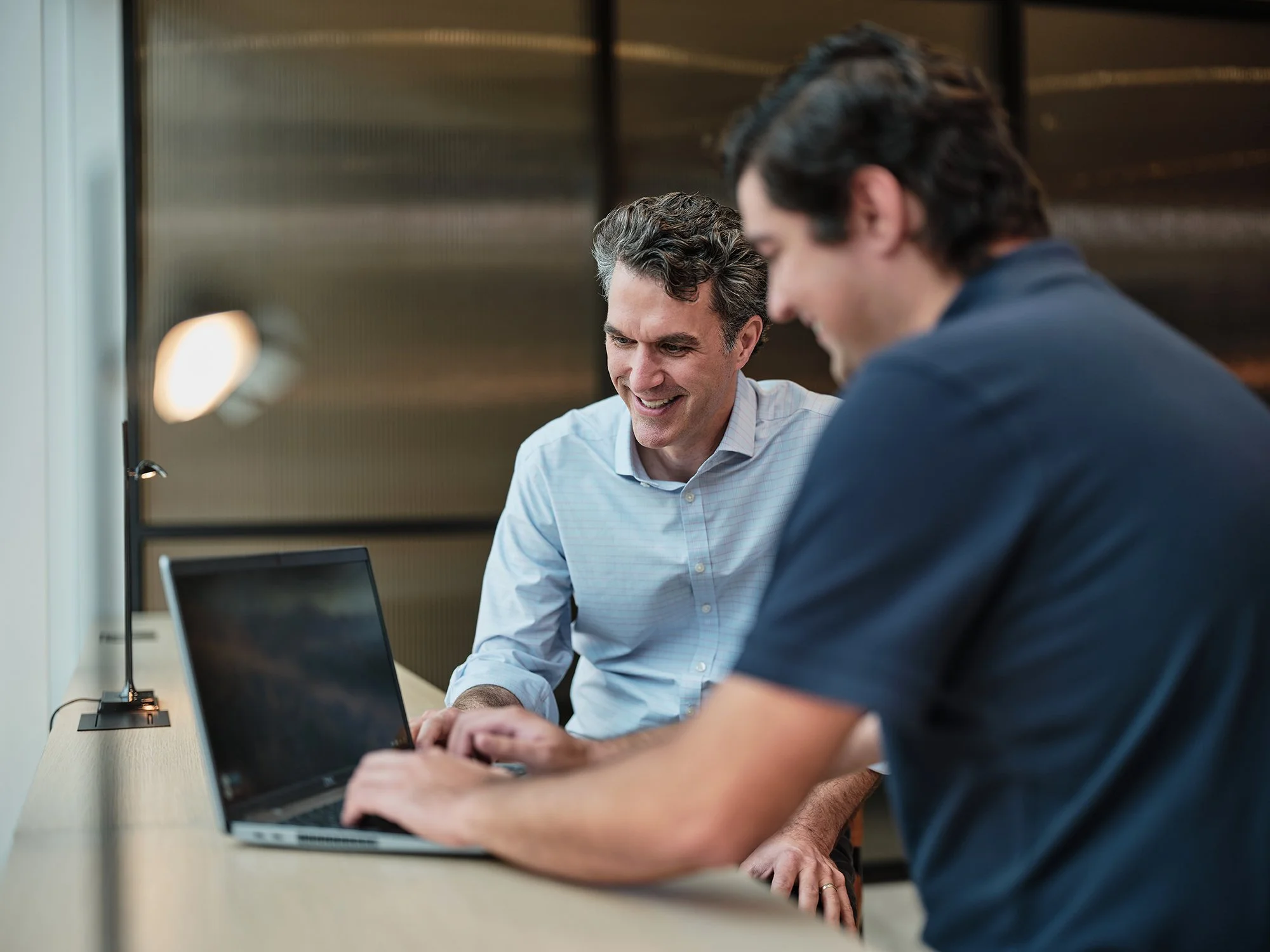 Two men sitting at a desk looking at a laptop screen, smiling and working together in an office setting.