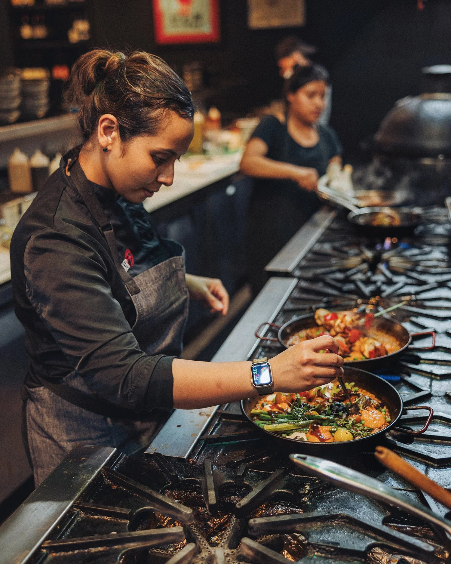 A female chef is cooking in a commercial kitchen, wearing a black jacket and apron, adding ingredients to a pan on a gas stove. Two women are working in the background.