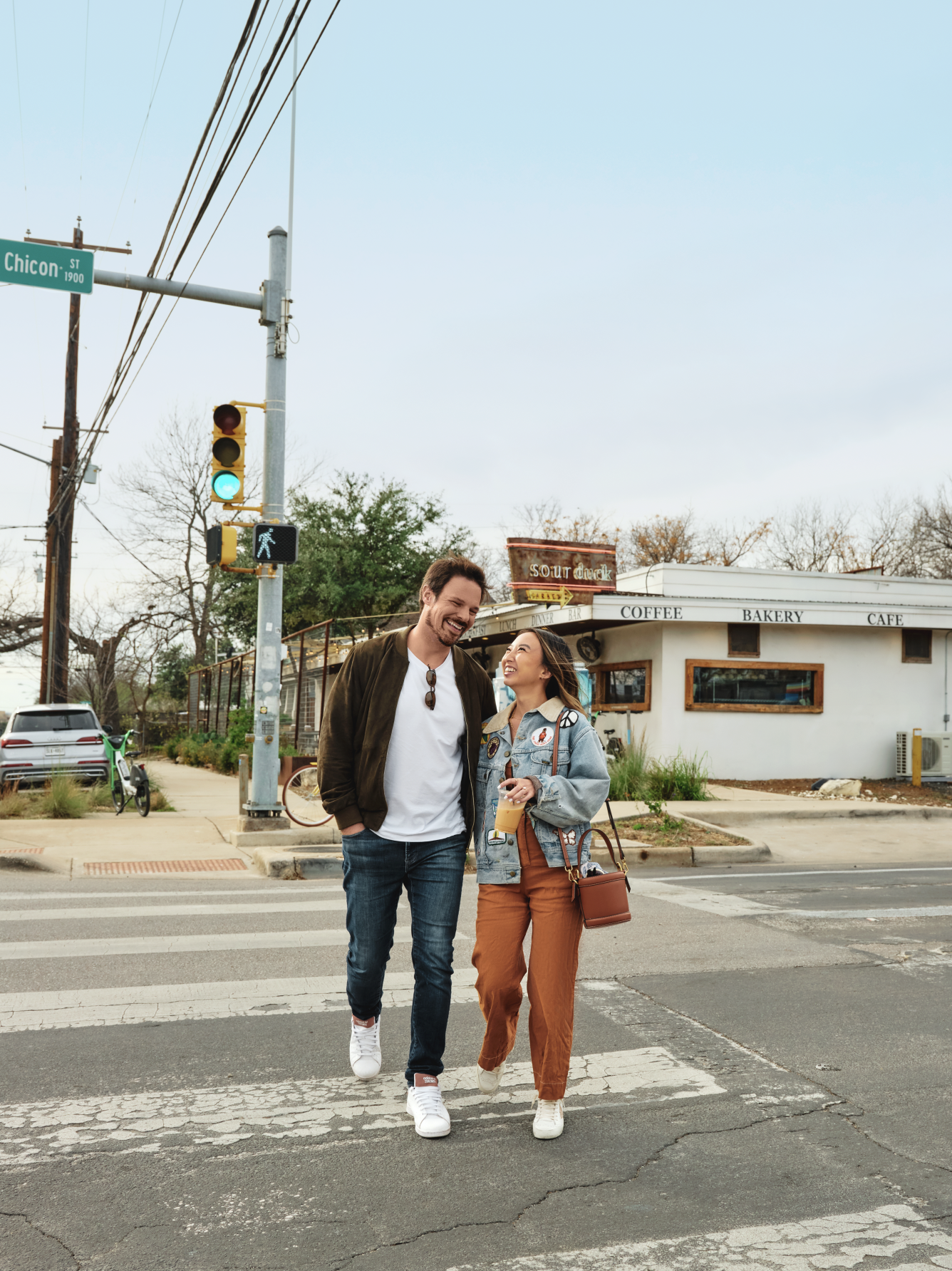 Young man and woman walking together across a crosswalk in an urban area, smiling and chatting, with a coffee shop in the background.