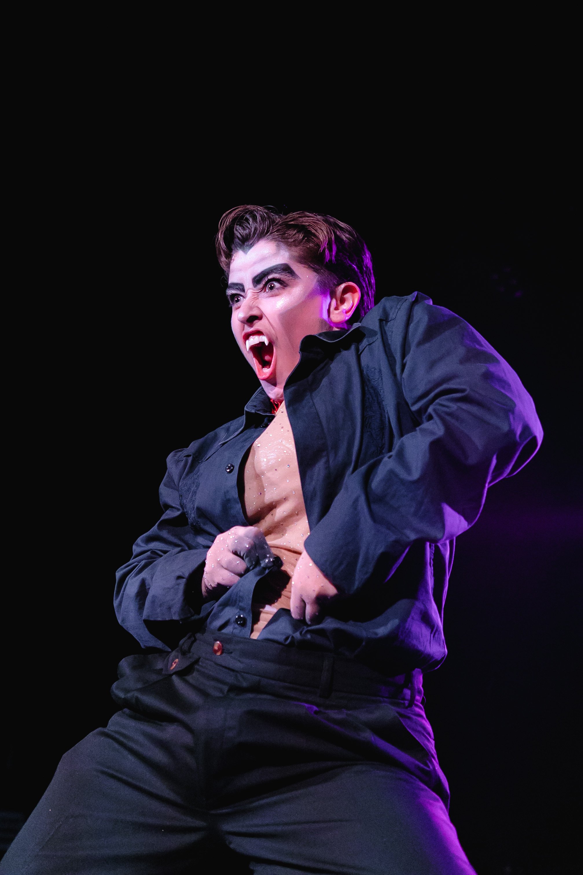 A woman with expressive face, dark hair, wearing a black jacket and beige top, performing on stage with bright lighting against a dark background.