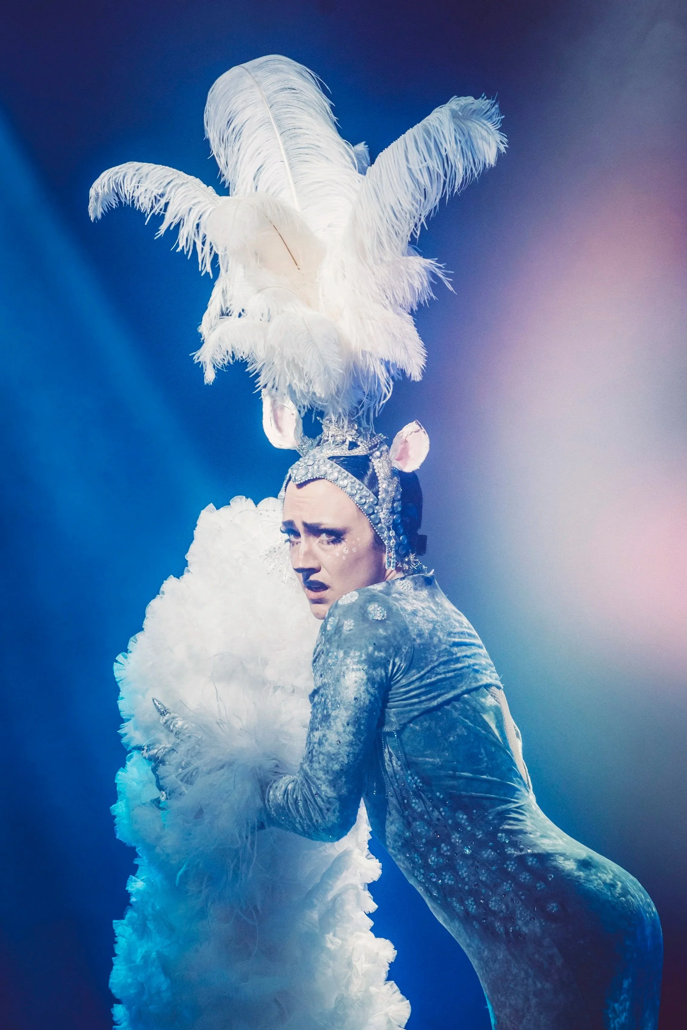 A performer dressed in an elaborate costume with white feathers and silver accents, wearing a headpiece with large white feathers, during a stage performance.