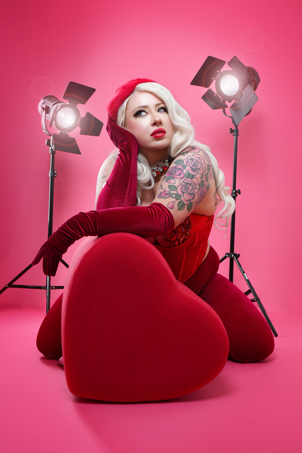 A woman with platinum blonde hair and tattoos on her arm poses on a pink background, sitting on a large red heart-shaped chair, surrounded by studio lights, wearing red gloves and a red dress.