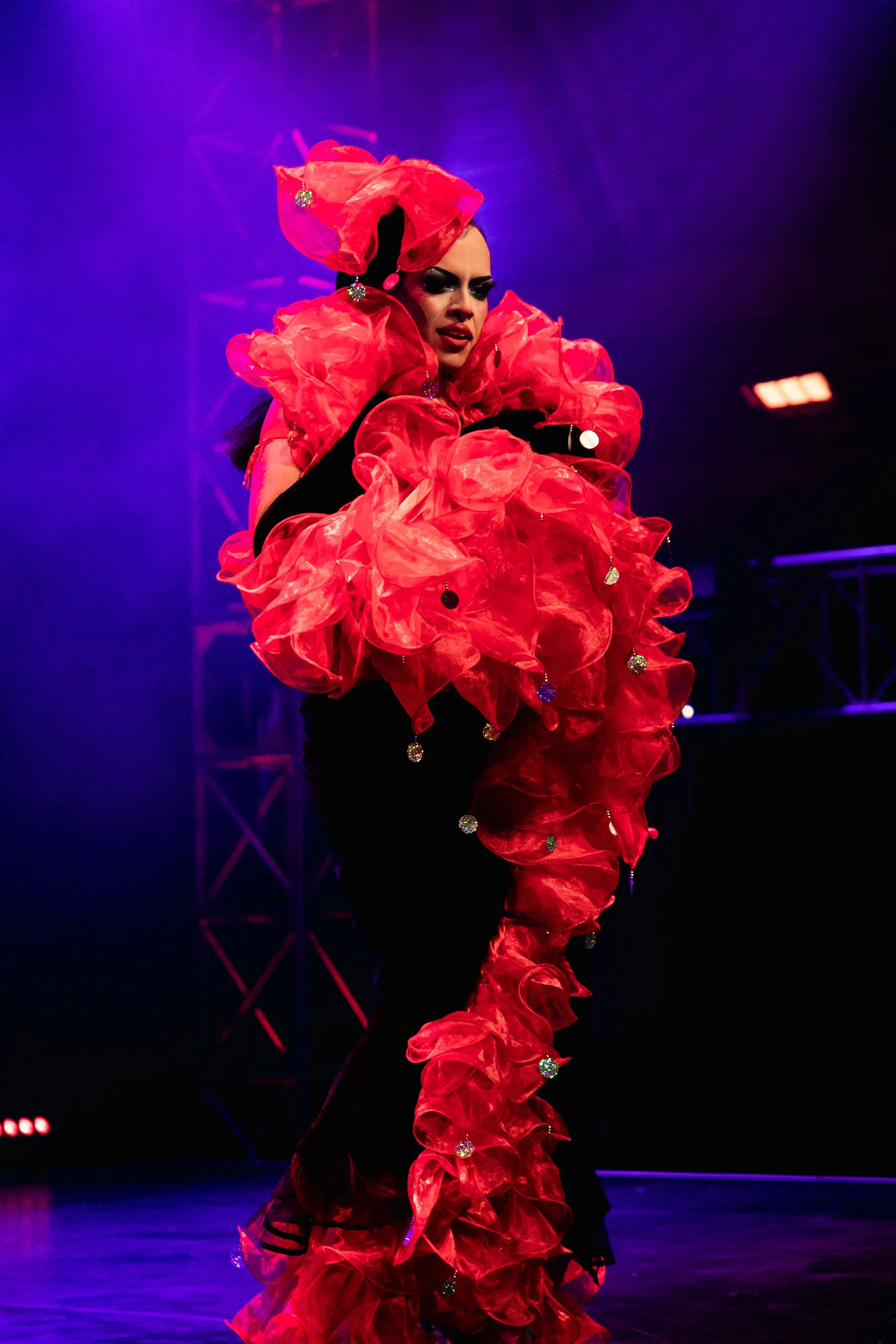 A performer on stage wearing a dramatic red and black ruffled costume with sparkly embellishments, under purple stage lighting.