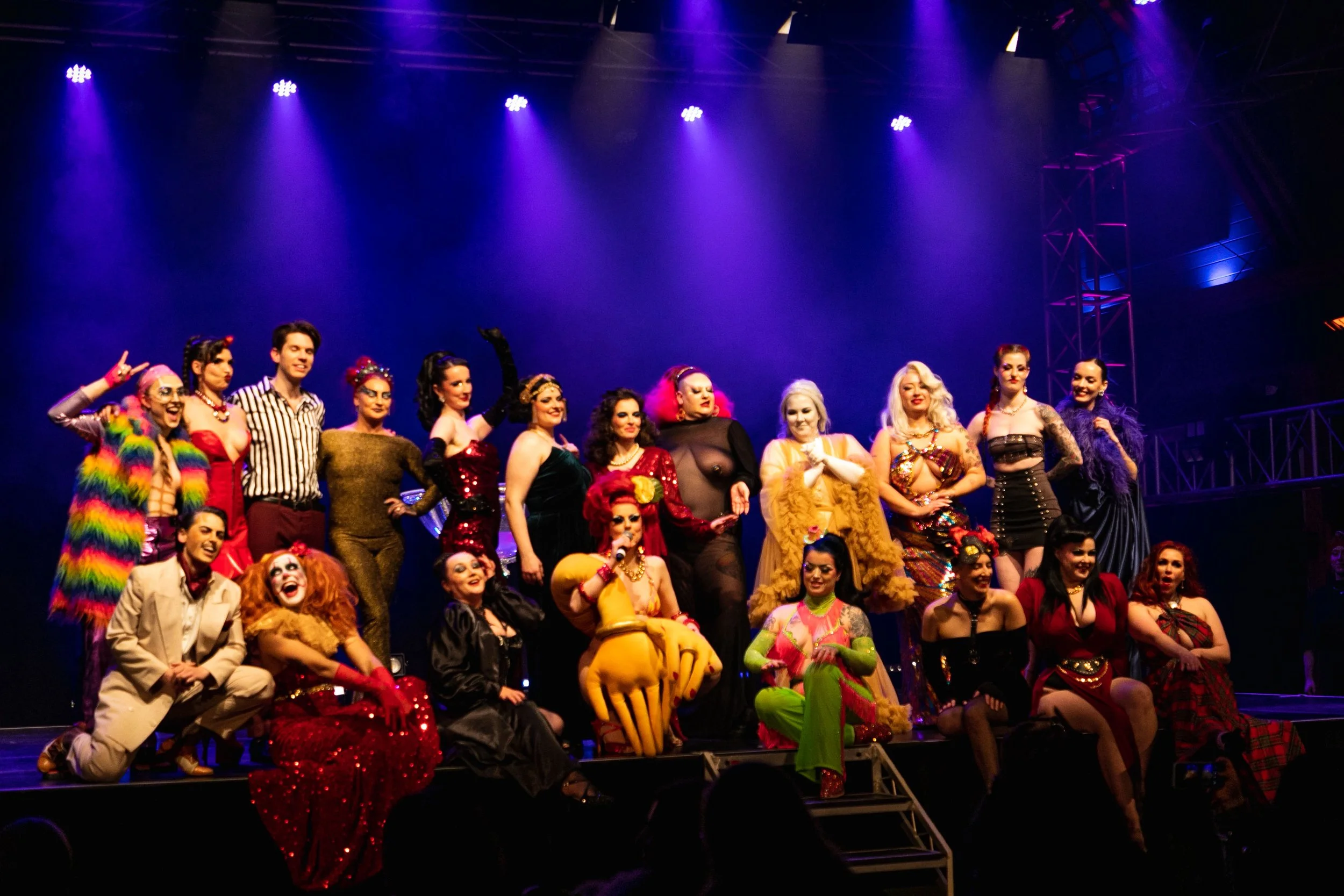 Group of performers in colorful costumes posing on stage after a show with purple stage lighting in the background.