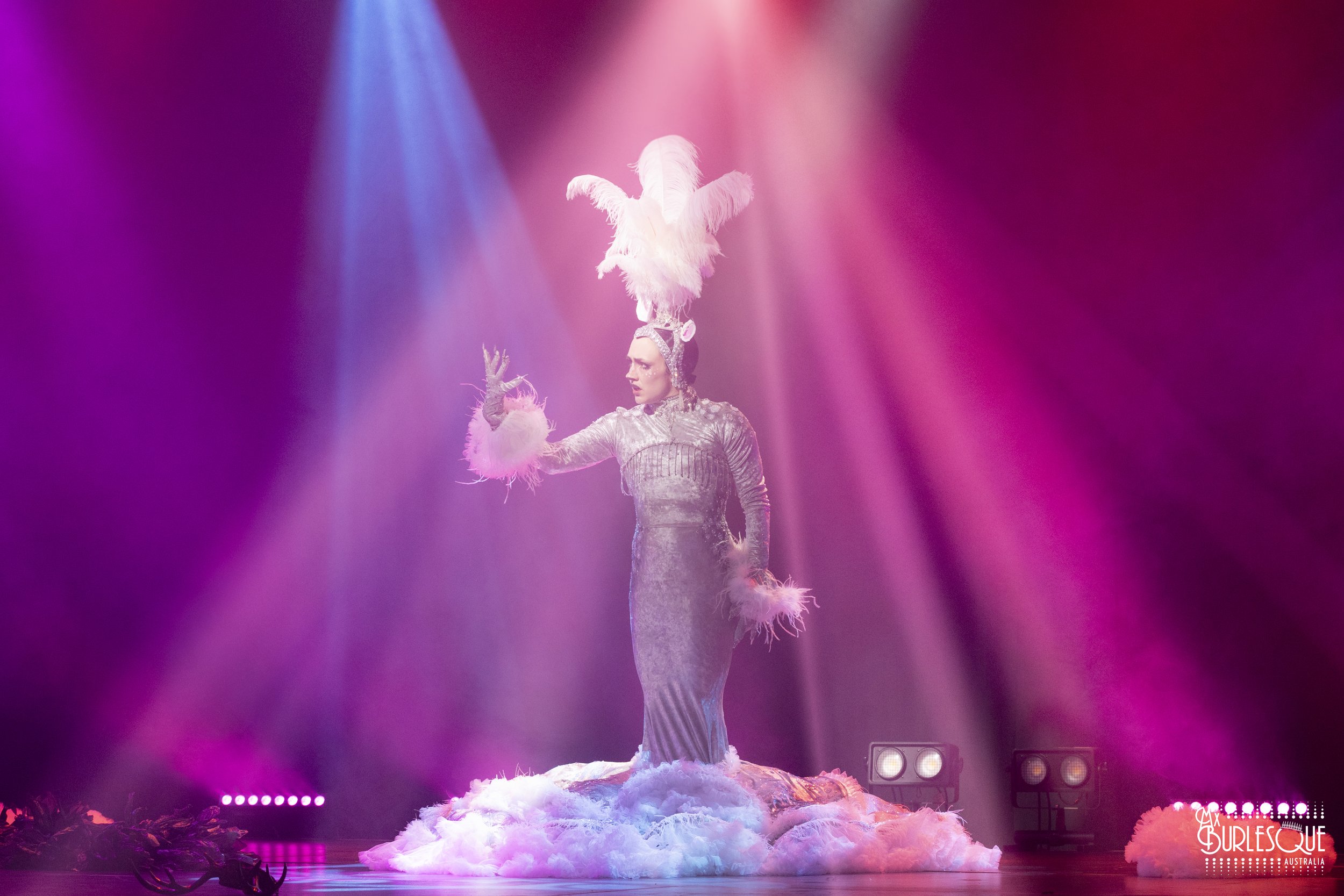 A performer dressed in a shiny silver costume with feathered cuffs and an elaborate headpiece with white feathers, standing on a cloud-like platform with pink, purple, and blue stage lighting.