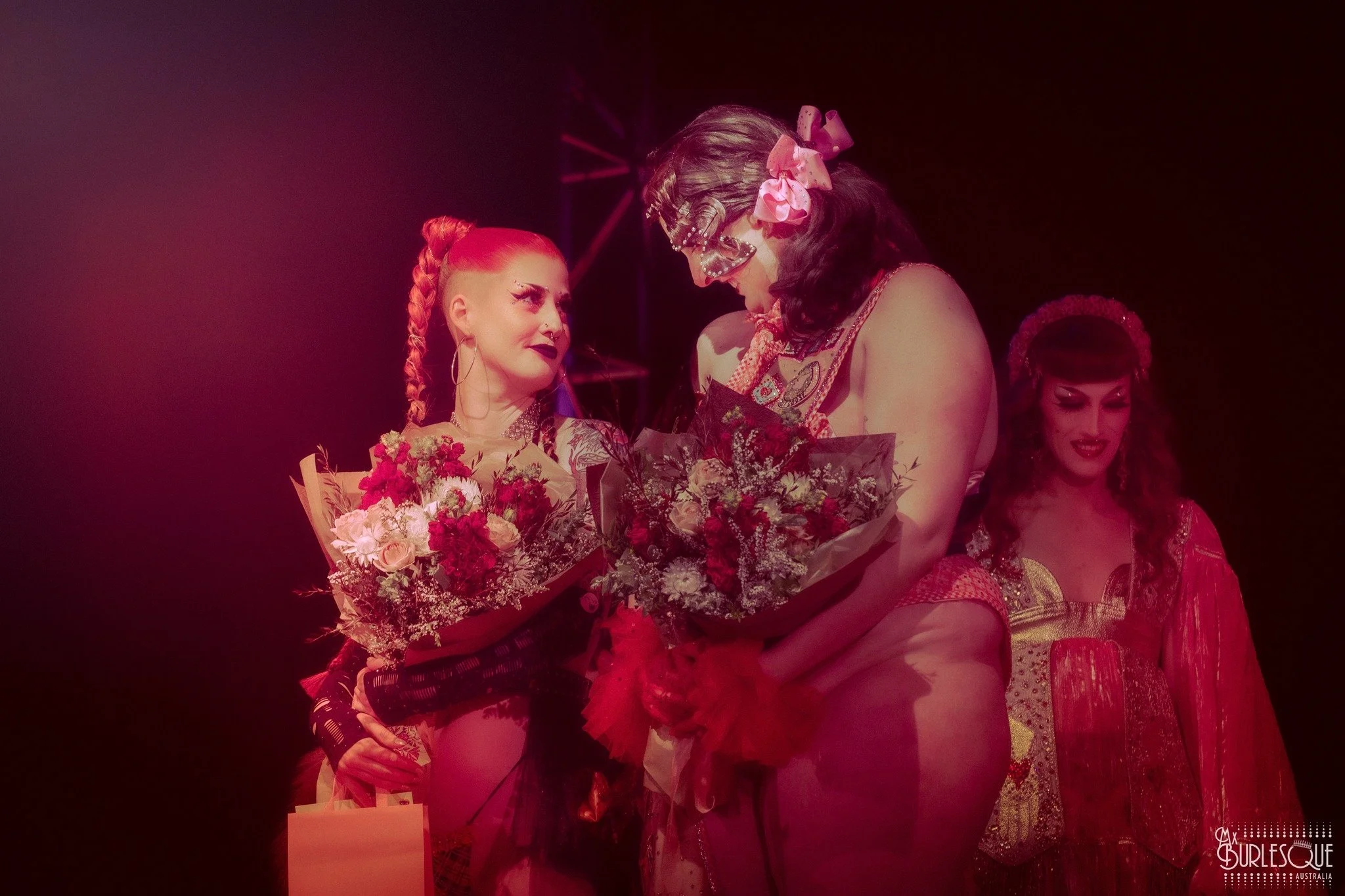 A performer dressed as a skeleton and wearing glasses with pink bows is presenting flowers to another performer with bright red hair styled in braids. Other performers are visible in the background on a stage.