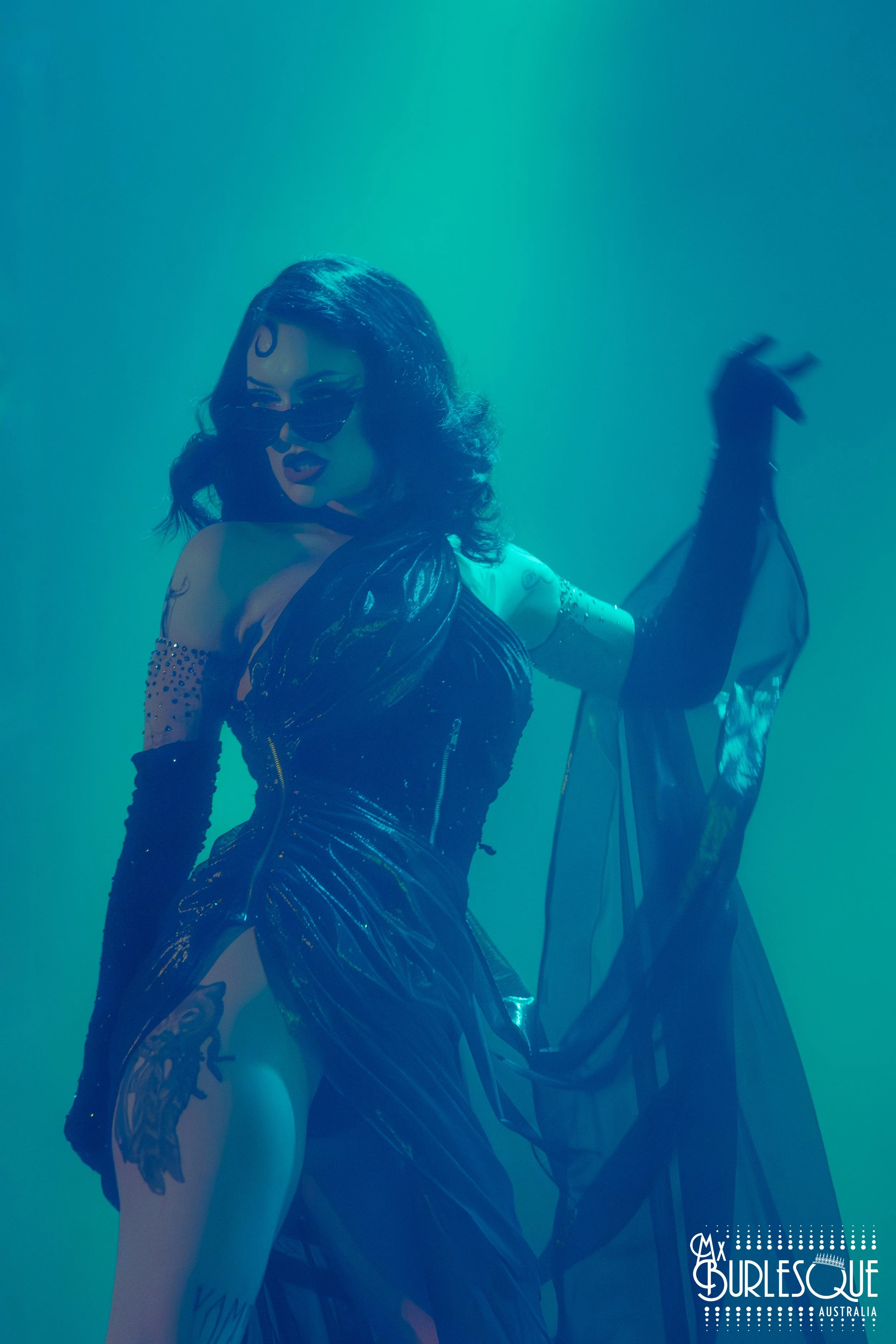 A woman with dark hair, wearing sunglasses, black gloves, and a shiny black dress, striking a pose against a teal background.