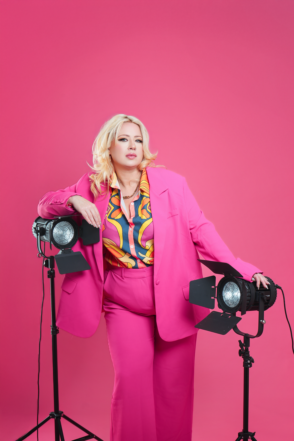 Woman with blonde hair in a pink suit standing in front of a pink background, next to studio lights.
