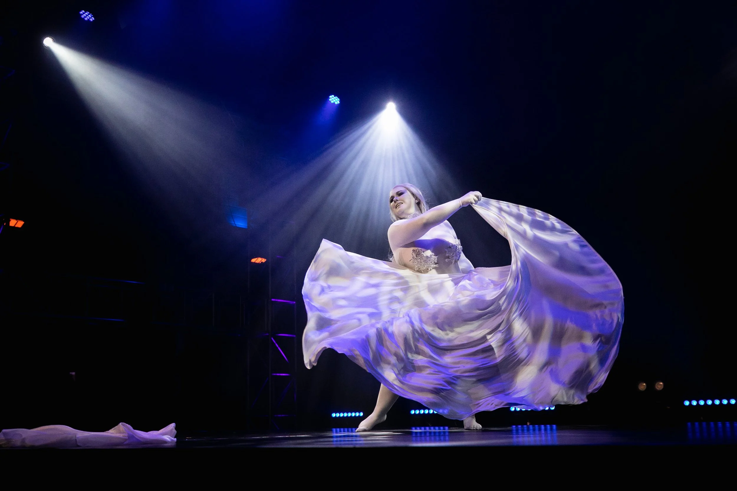 A woman twirling in a flowing, purple and white dress on a dark stage with bright stage lights overhead.