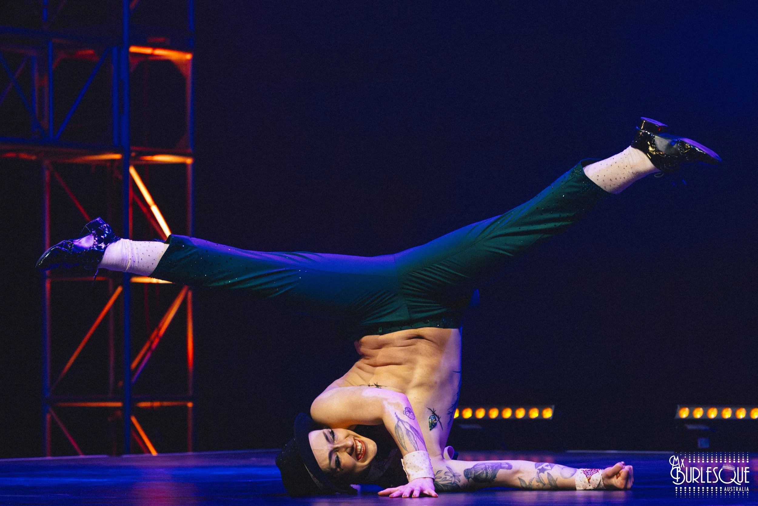 A perfor performing a contortion pose on stage, balancing with one hand while their legs are extended upward and horizontally, smiling and wearing black shoes, tattoos visible on her arms, with stage lighting and scaffolding in the background.