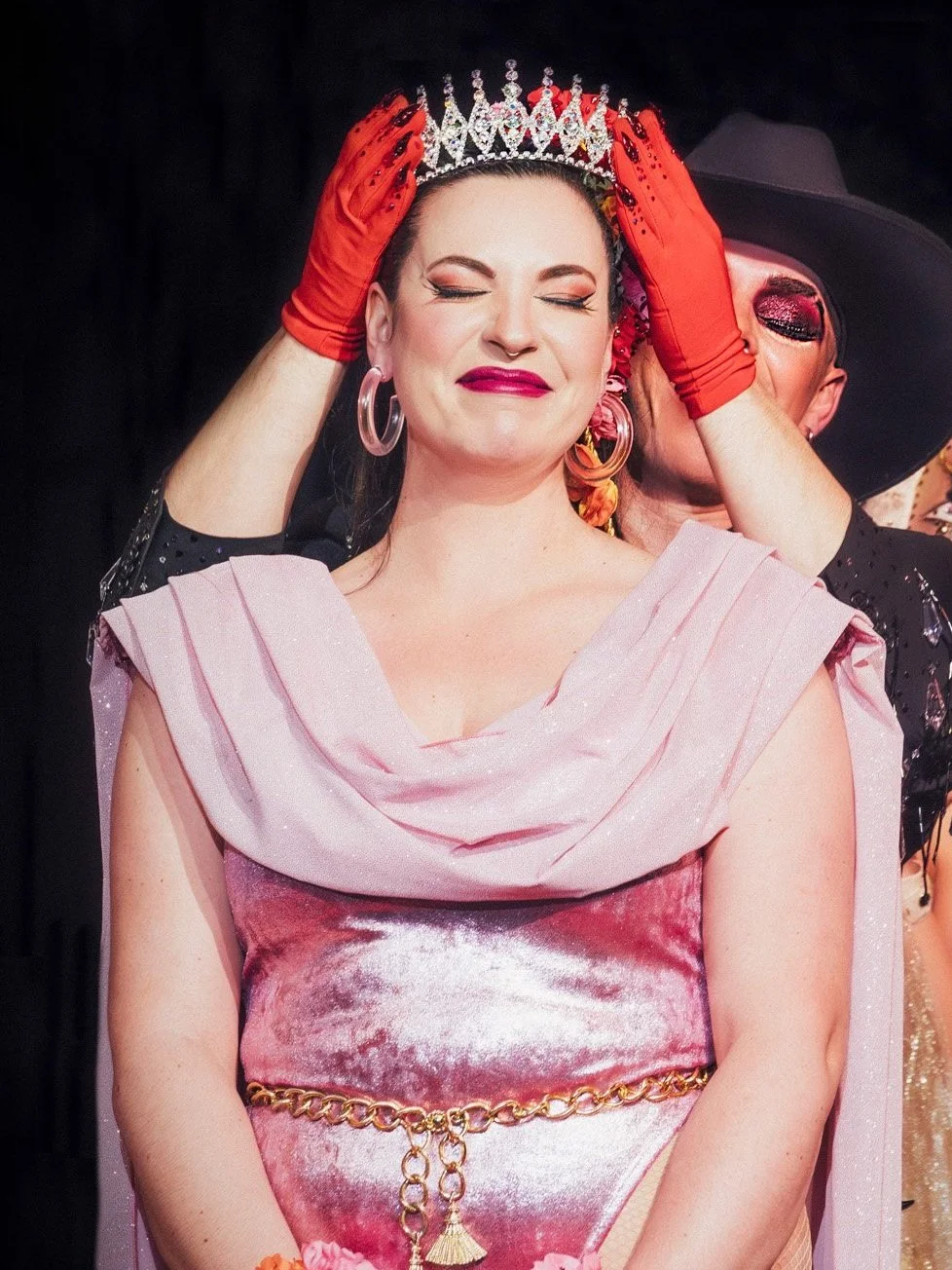A woman dressed in a glamorous pink outfit with a gold chain belt, wearing large hoop earrings and makeup. A performer places a crown on her head while wearing red gloves. The scene appears to be a theatrical or pageant event.