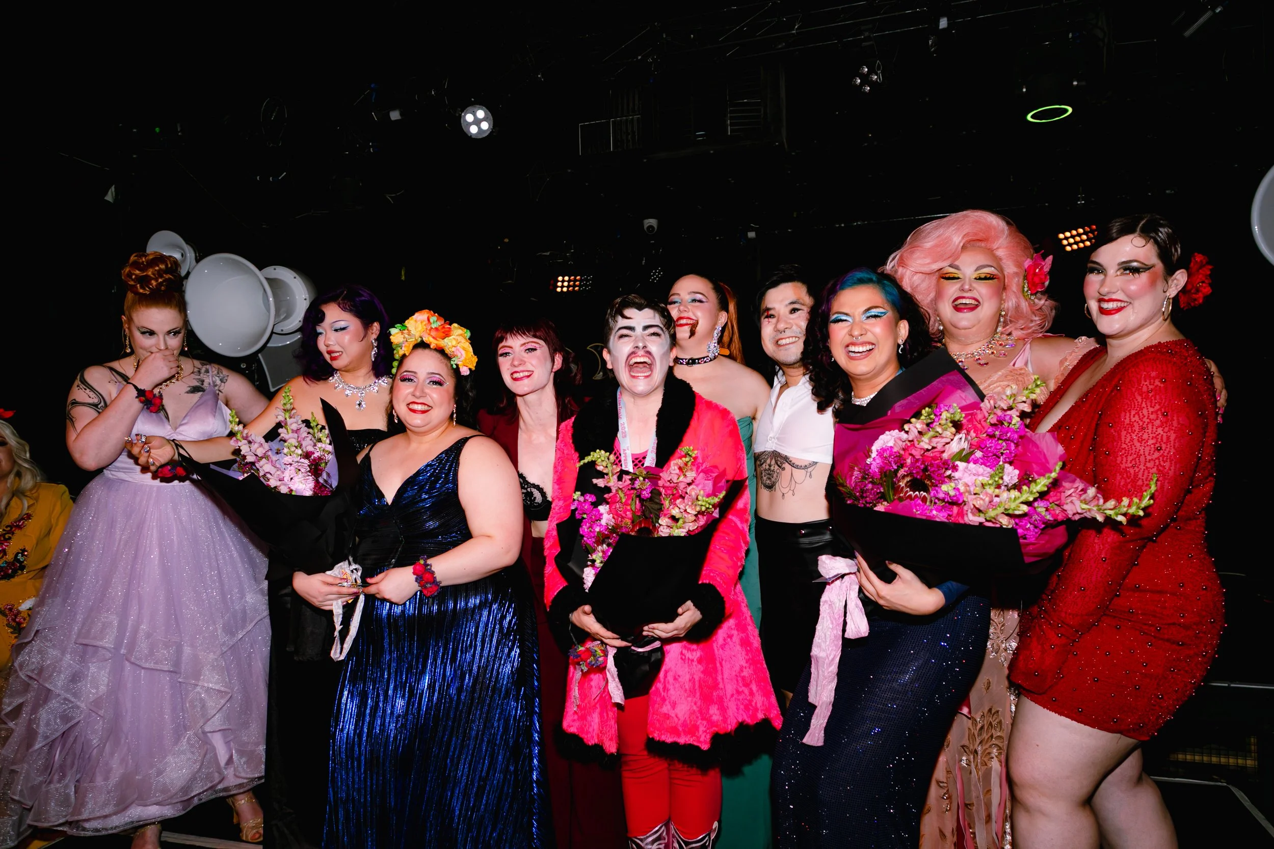 Group of performers in colorful costumes and makeup, holding bouquets, on stage at a celebration or performance event.