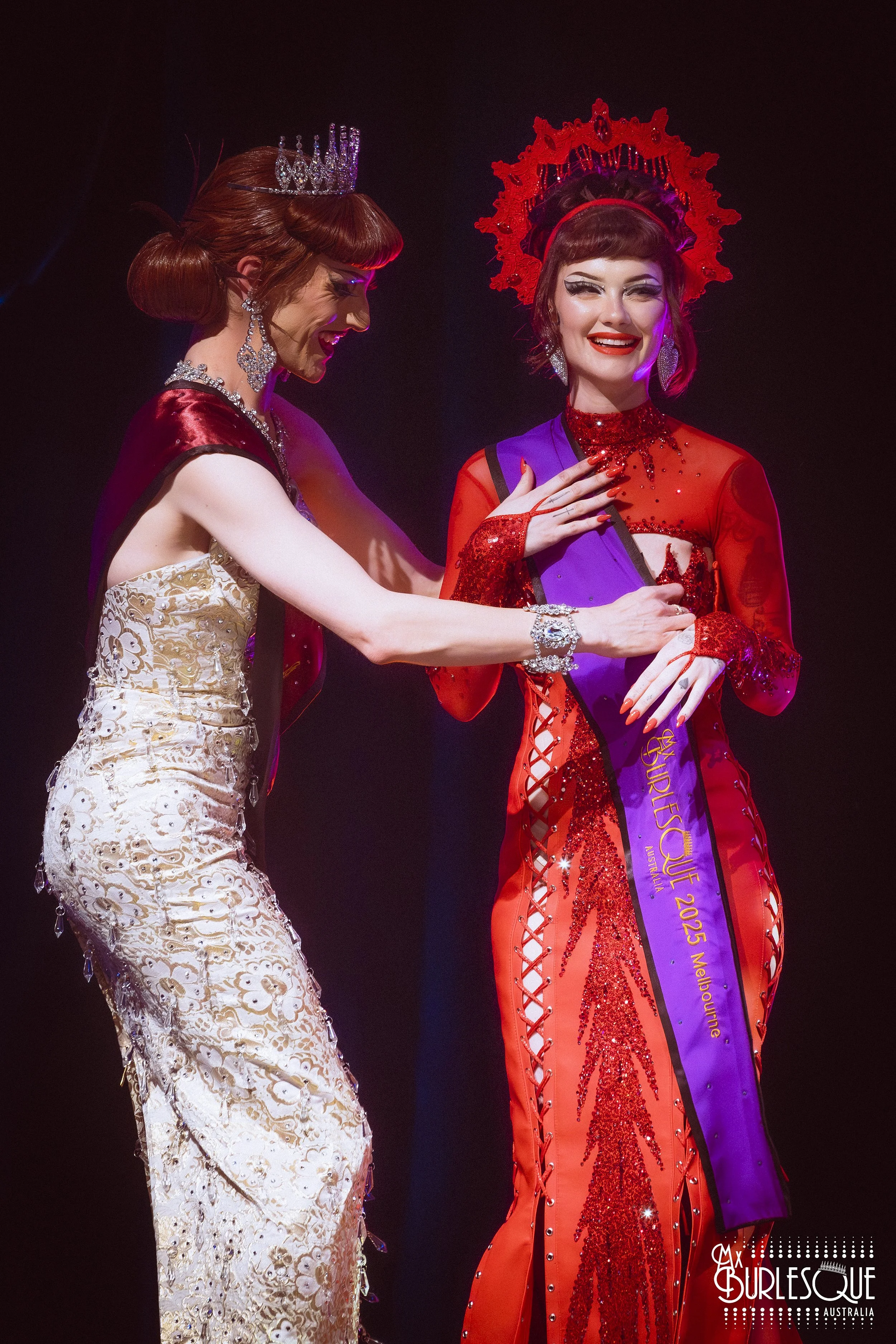 Two women on stage, one wearing a white and gold gown with a tiara, and the other wearing a red dress with sequins and a sash that reads "Ms. Beautiful 2025 Melbourne," exchanging an object with smiles.