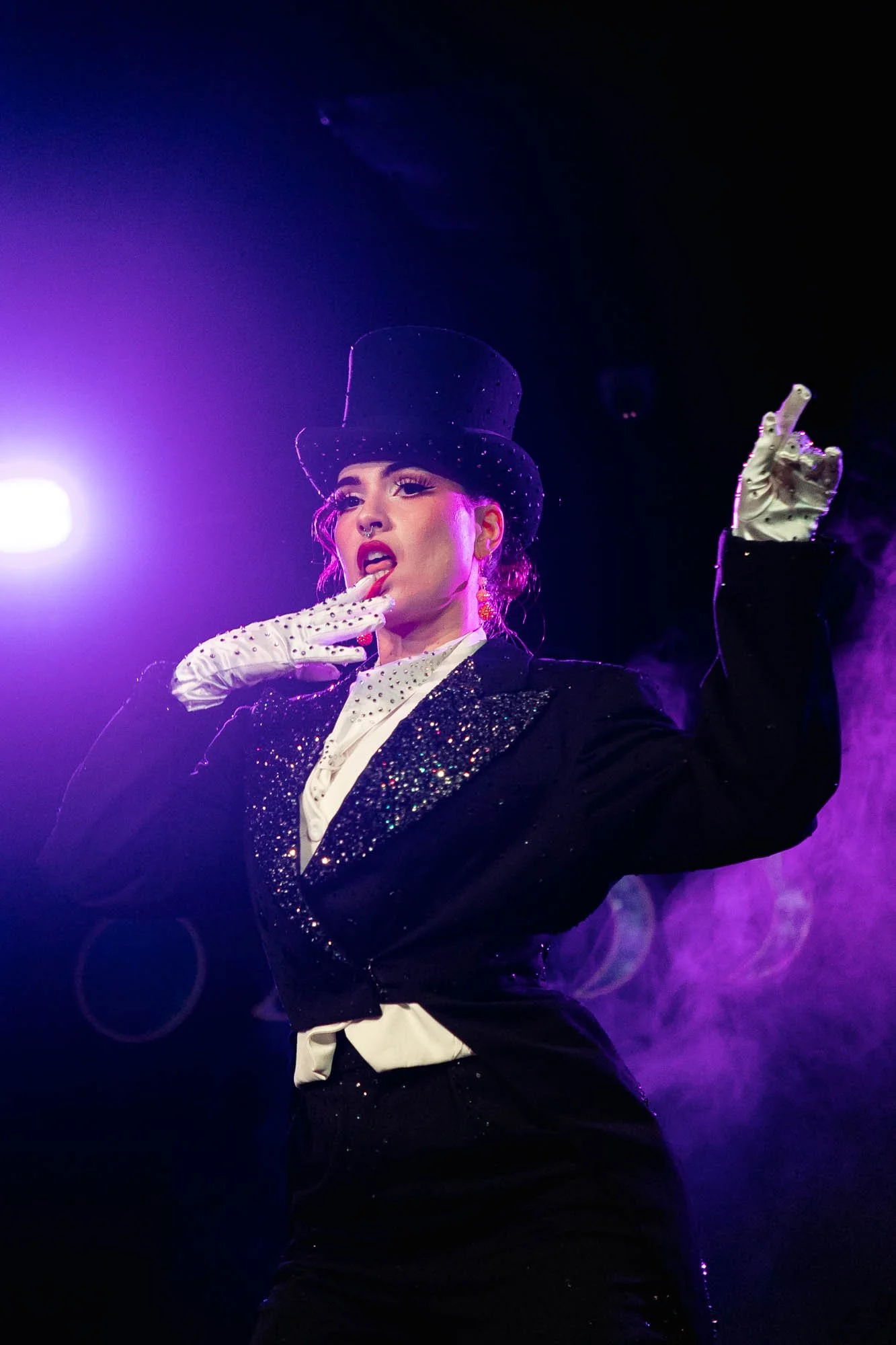 Performer on stage wearing a black top hat and a black sequined jacket, with white gloves, striking a pose with one hand near her face and the other raised, under purple stage lighting.
