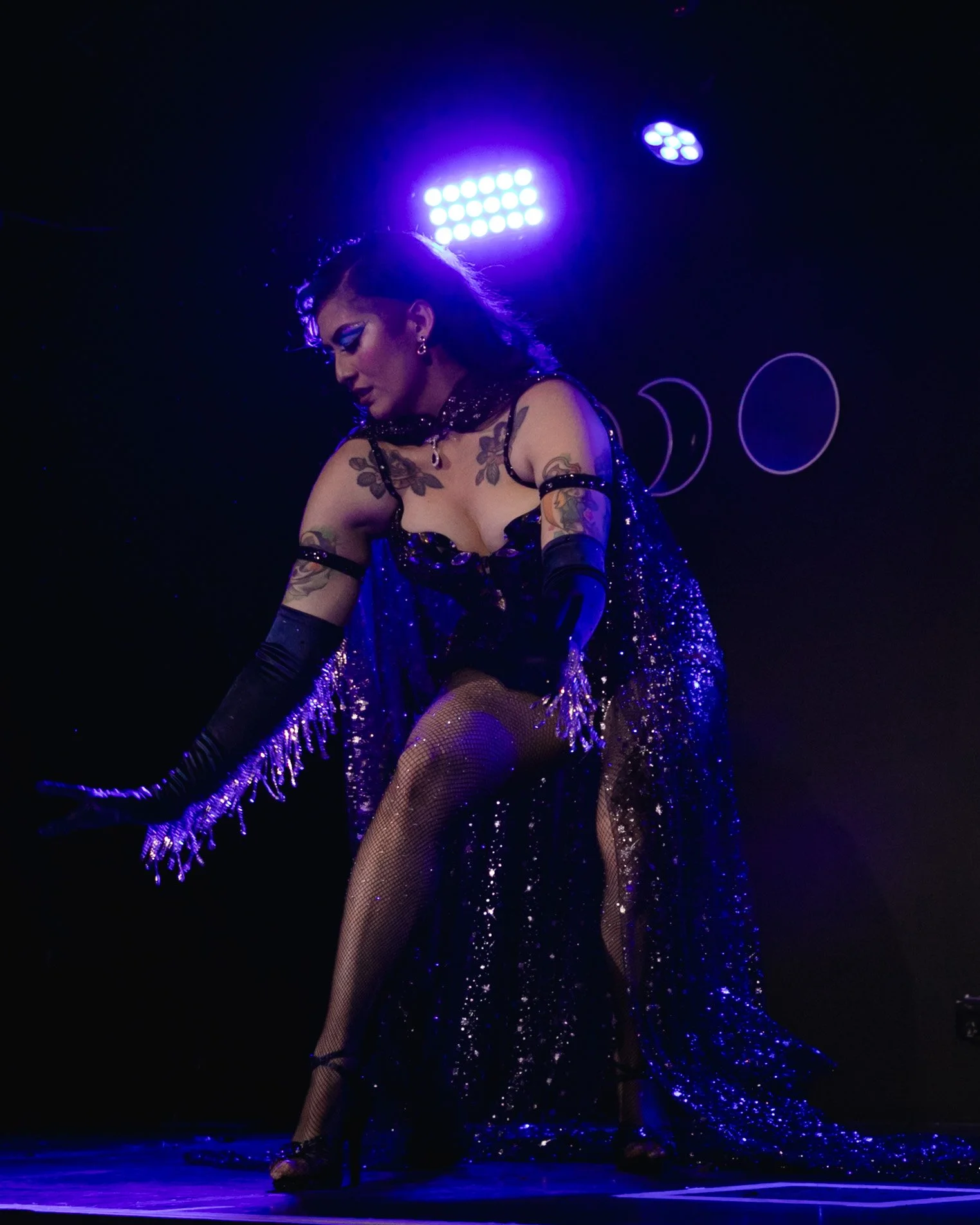 A woman dressed in a sparkly black and purple costume performing on stage with purple lights and a dark background.