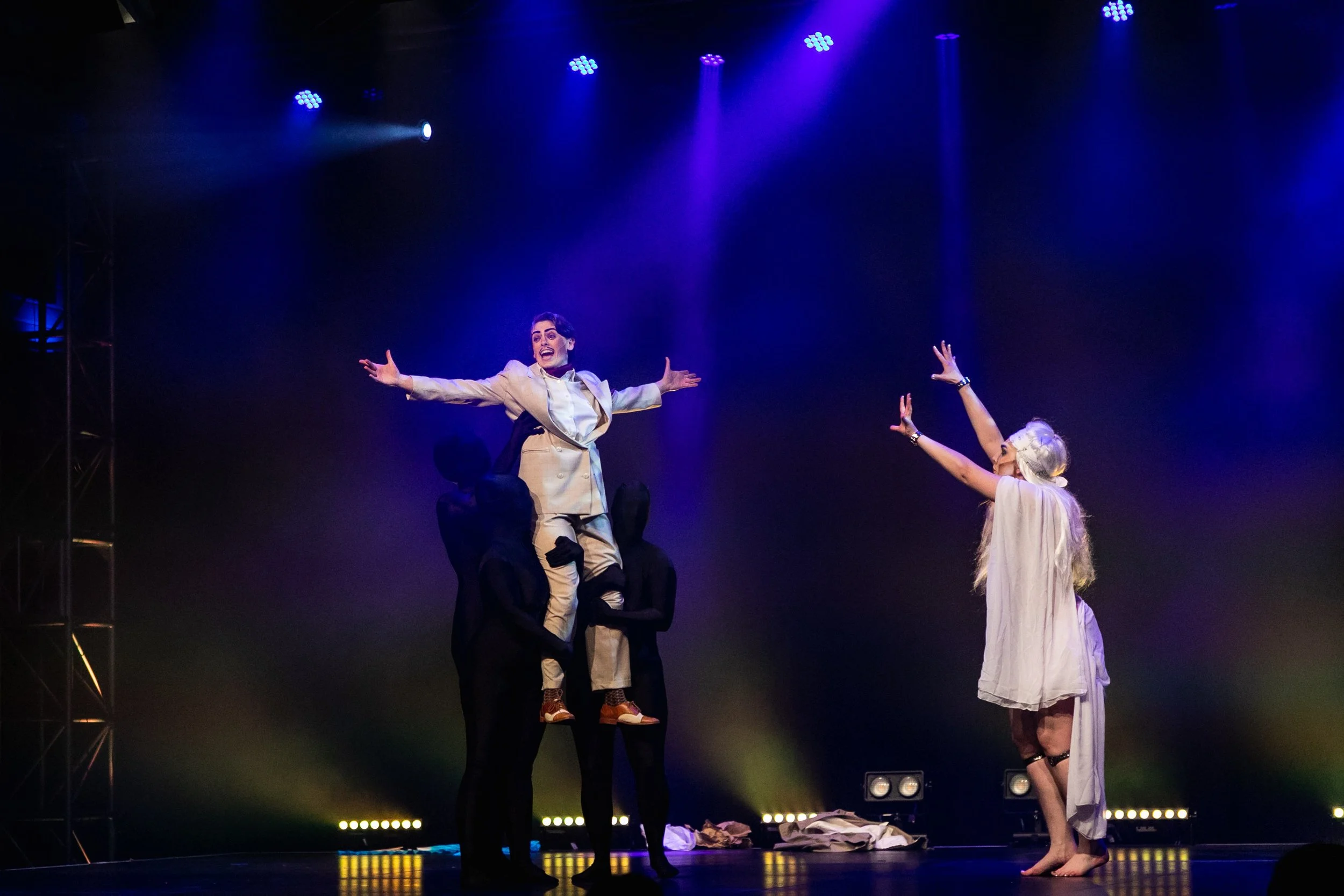 A theatrical performance with a person being lifted by four others on stage, with one woman in front reaching out towards the person with outstretched arms, under purple stage lighting.