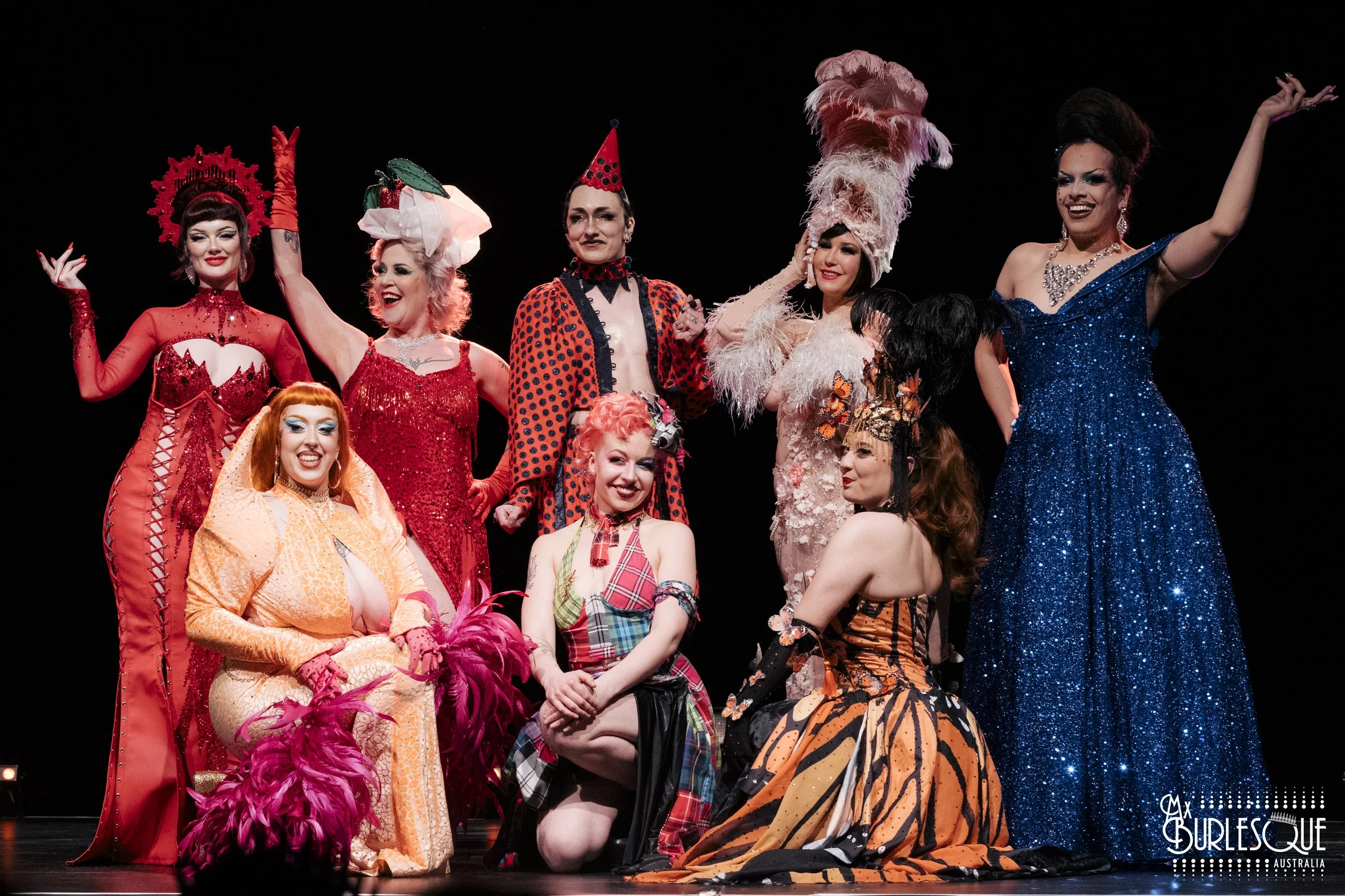 Ten diverse performers in colorful, elaborate costumes pose on a stage against a black background, possibly in theatrical or burlesque performance.