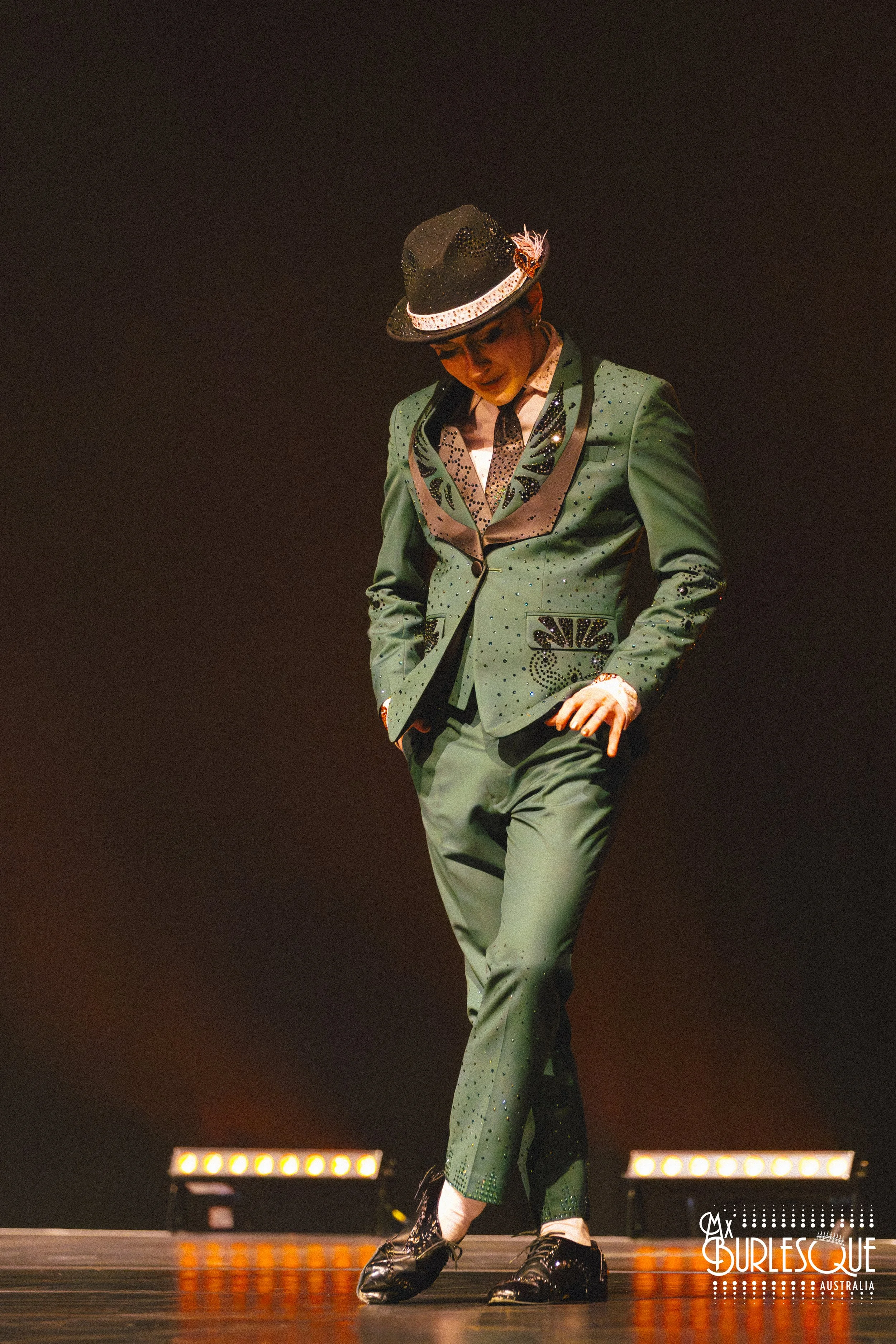 Performer dressed in a green, sequined suit with black and white accents, wearing a black hat with sequins and a feather, standing on stage with one hand in pocket, under orange stage lights.