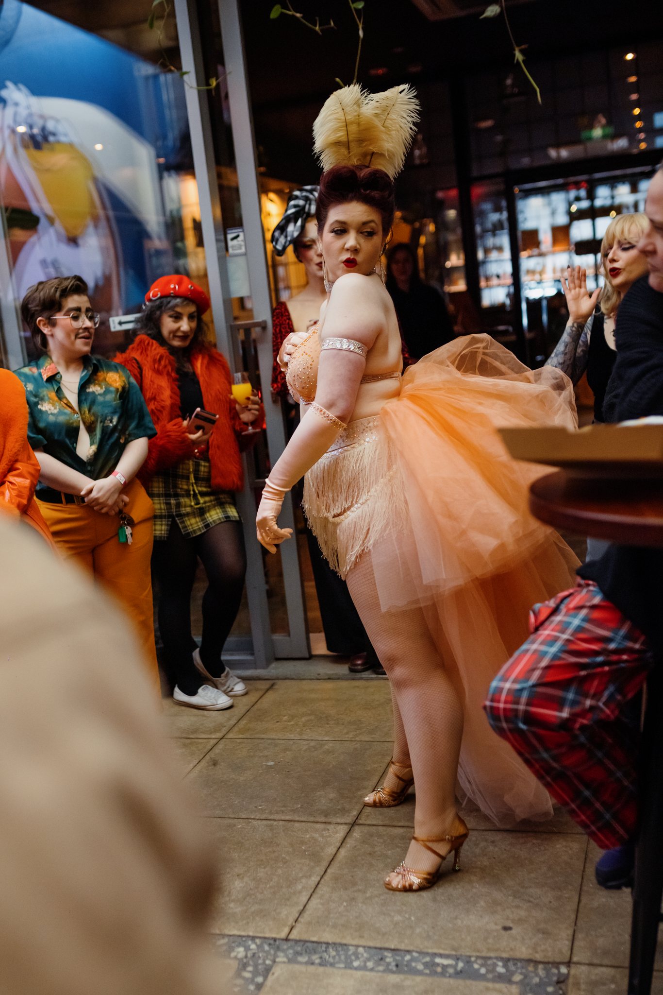 A woman dressed in a peach-colored ball gown with tulle fabric and wearing high-heeled shoes is dancing or posing, surrounded by a group of women dressed in colorful attire with one woman in a red hat and another in an orange furry coat. The scene appears to be in a restaurant or bar with glass walls and warm lighting.
