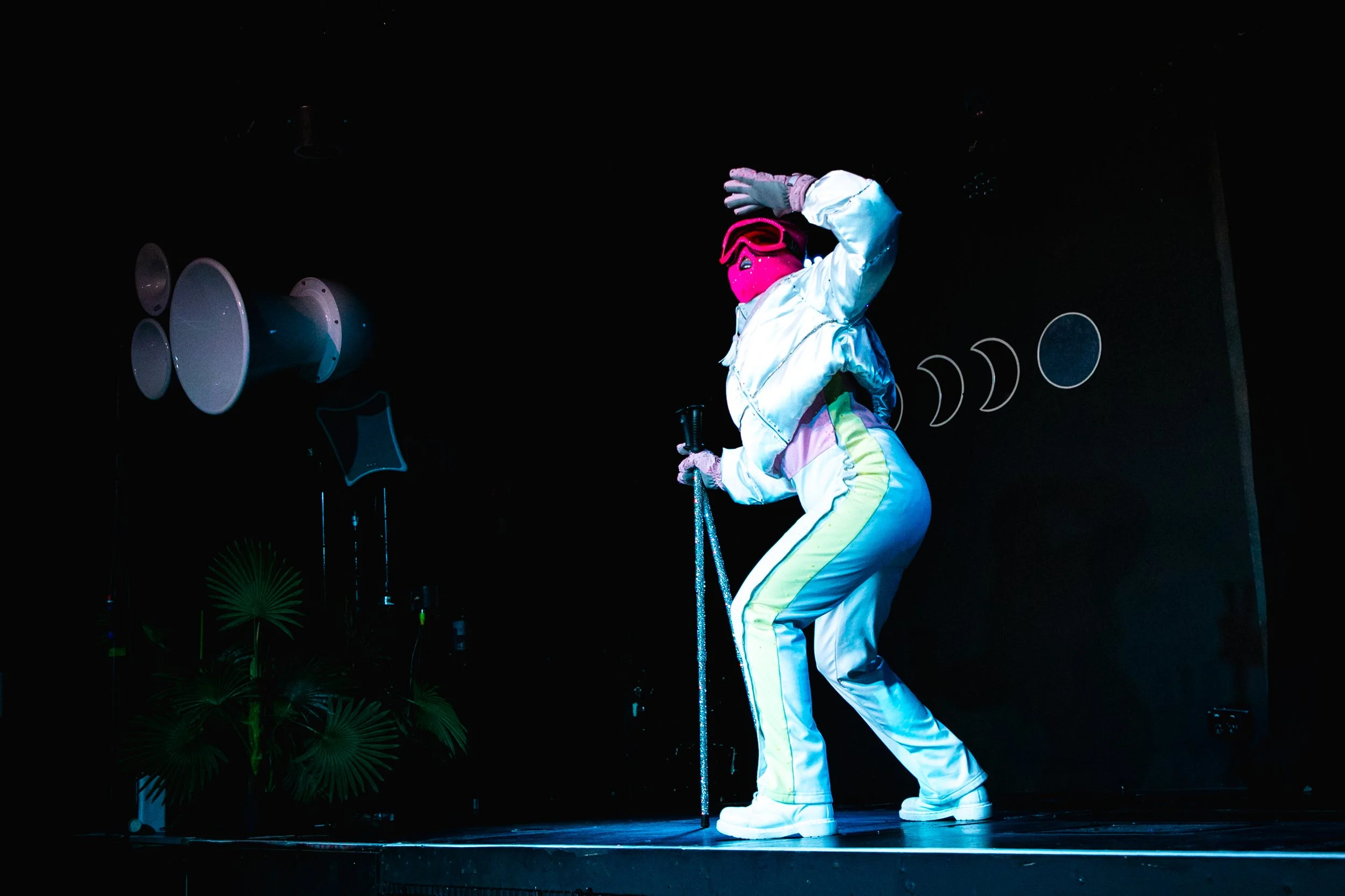 Performer dressed in shiny white and yellow outfit with pink mask and goggles, holding a cane, on stage with dark background and stage lighting.