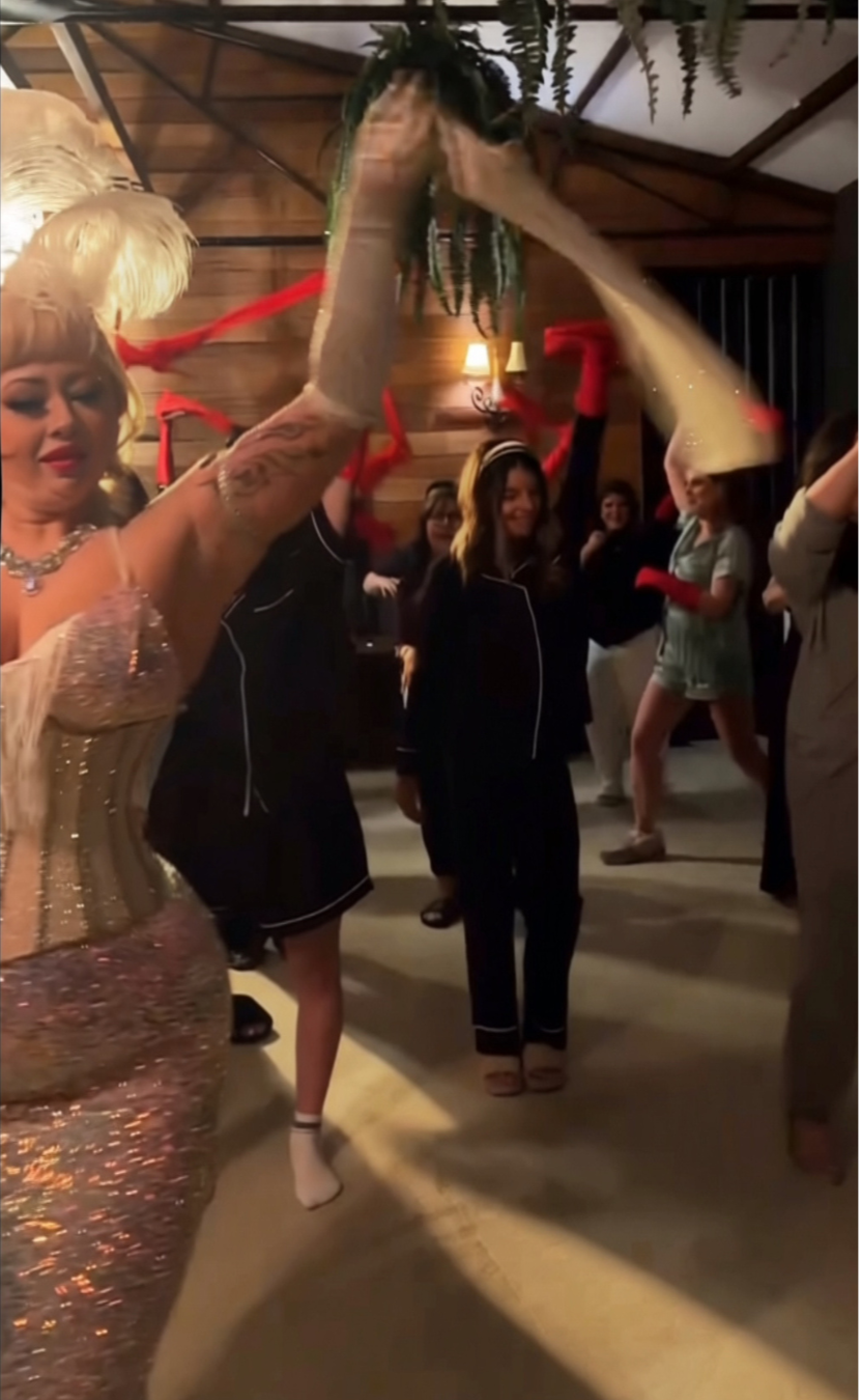 Burlesque hens party Melbourne, with people dancing, with a person dressed in a glamorous gold gown with a feathered headpiece and a person in black pajamas with a headband, holding a leafy plant above their head.