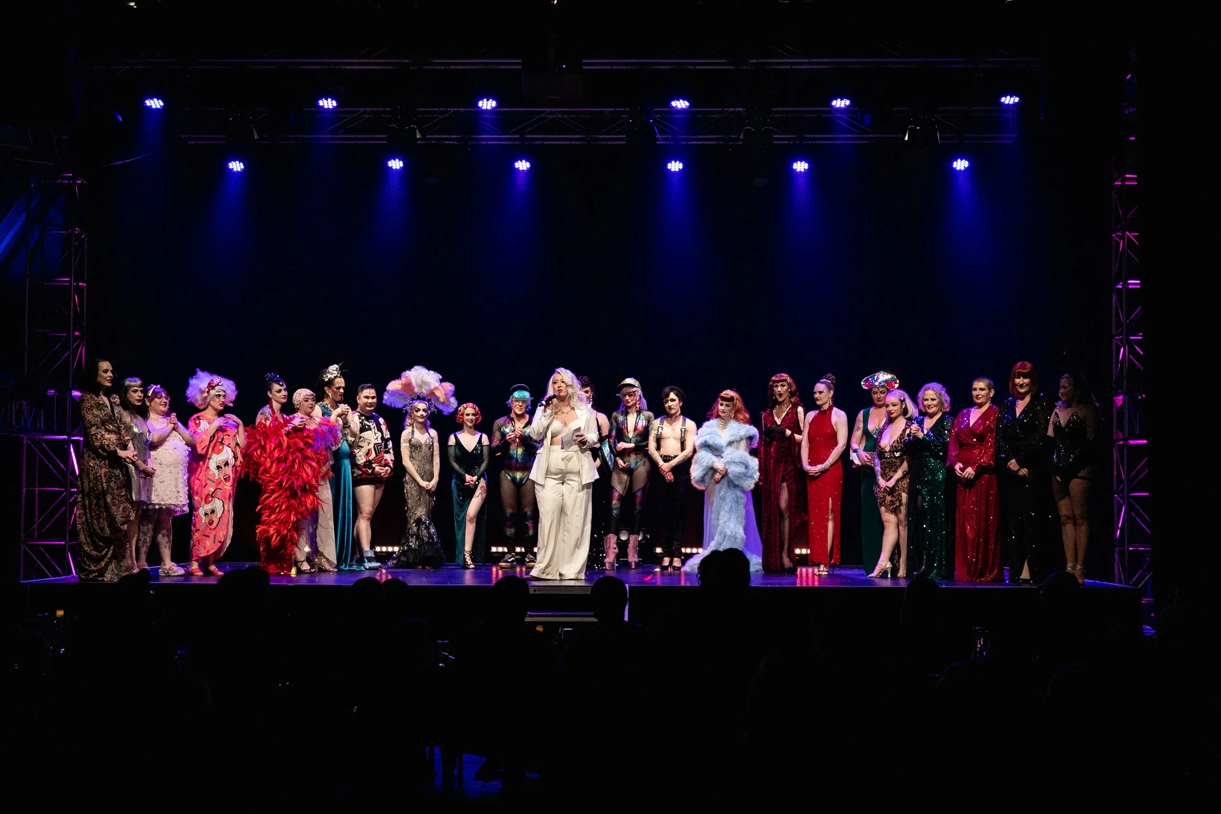 A stage performance featuring a diverse group of performers dressed in colorful, elaborate costumes, with blue stage lighting and an audience silhouette in the foreground.