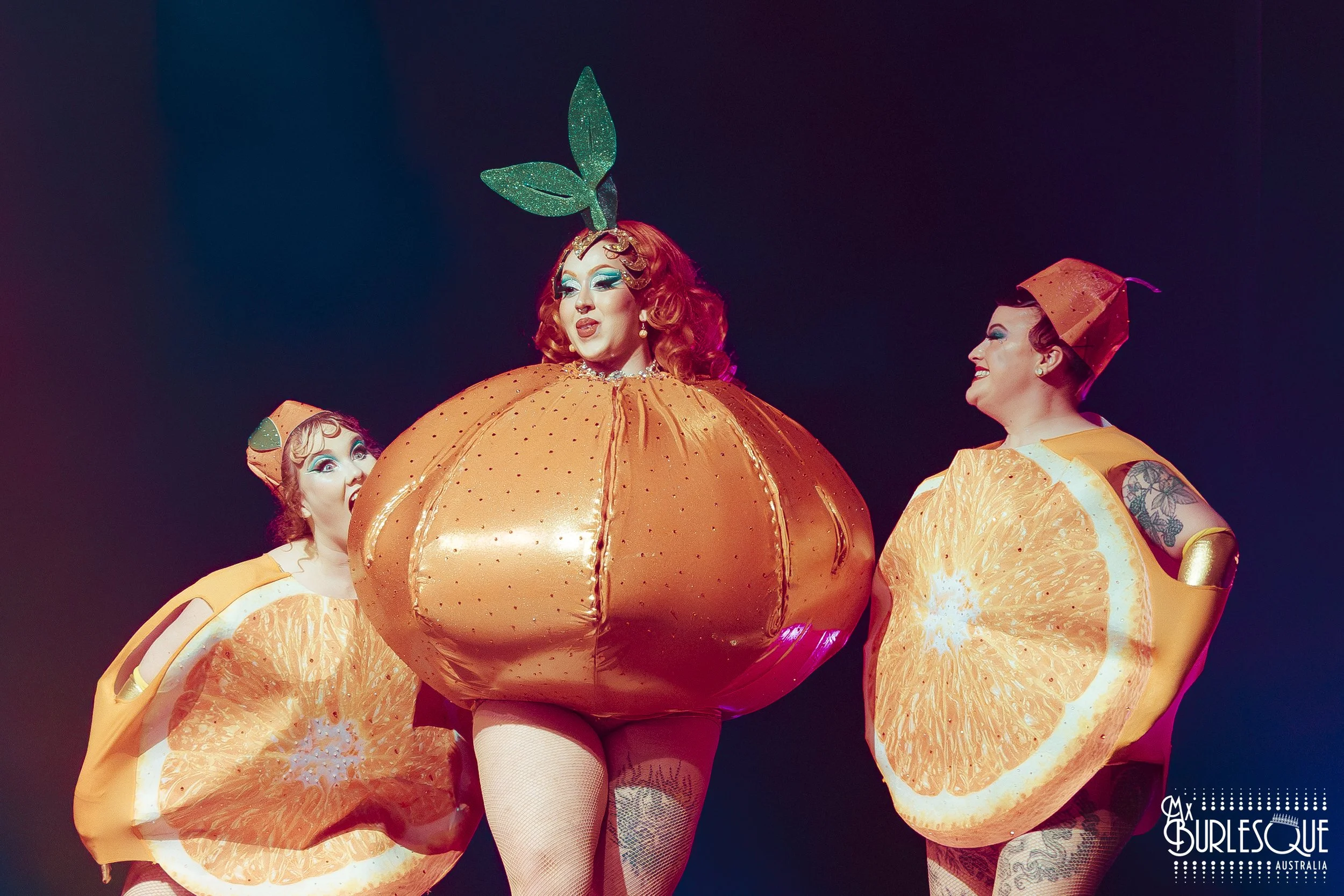 Three performers dressed as oranges and a person dressed as a pumpkin with a green leaf hat, on stage with dark background
