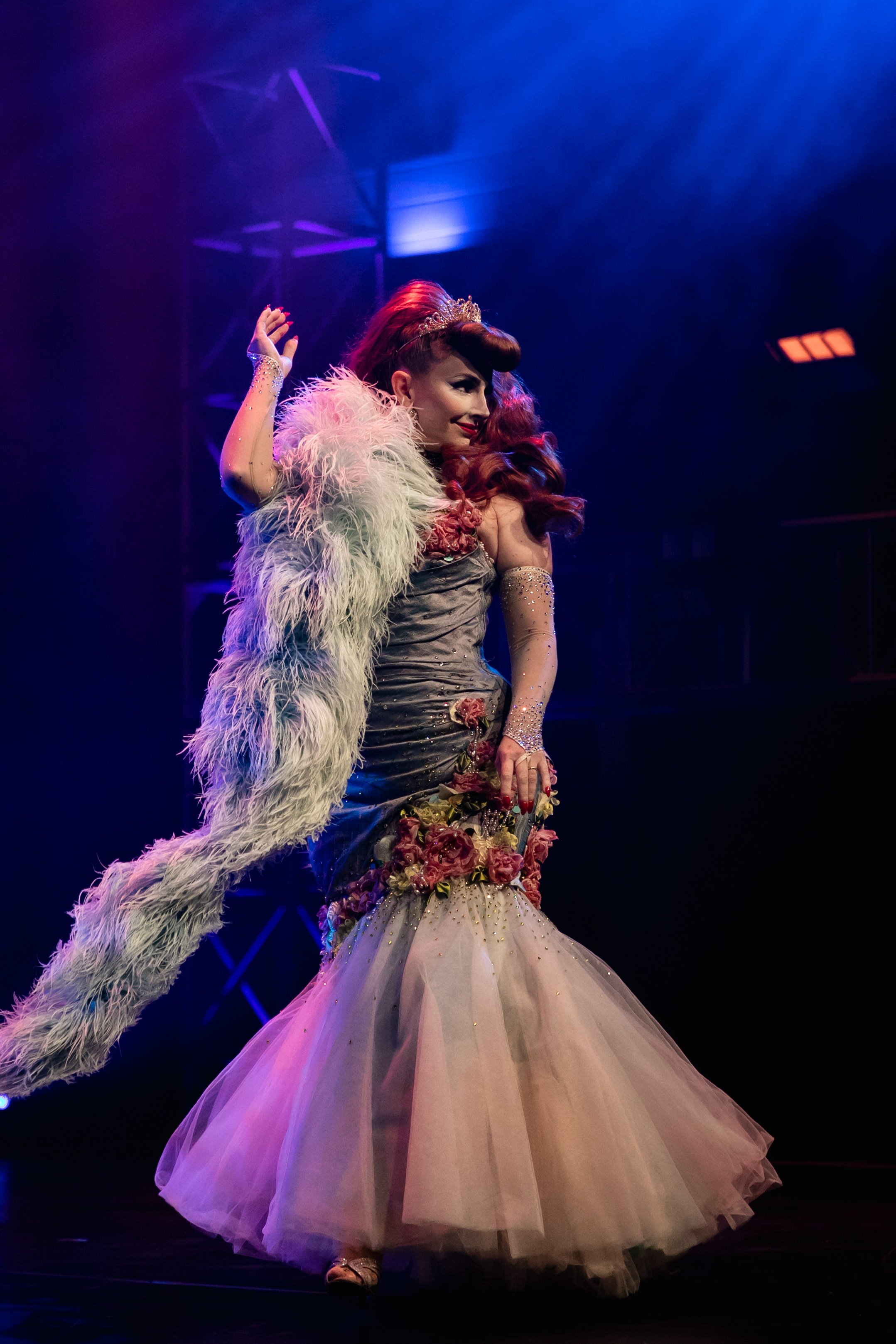A woman on stage wearing a elaborate gown with floral details and a feathered cape, with vintage hair and makeup, under dramatic lighting.