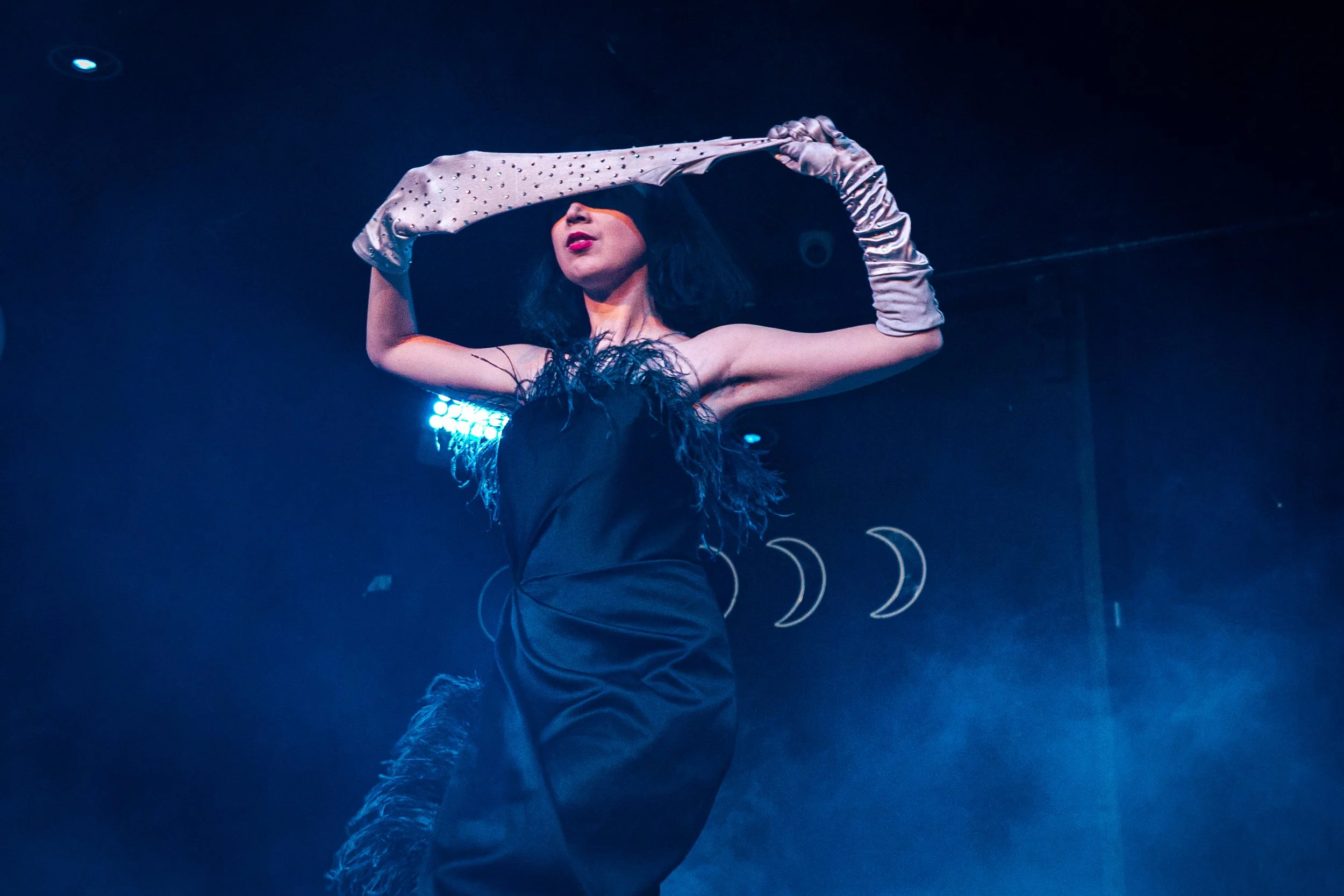 A woman on stage wearing black dress with feather details, long beige gloves, and feathered boots, striking a pose with her arm raised above her head.