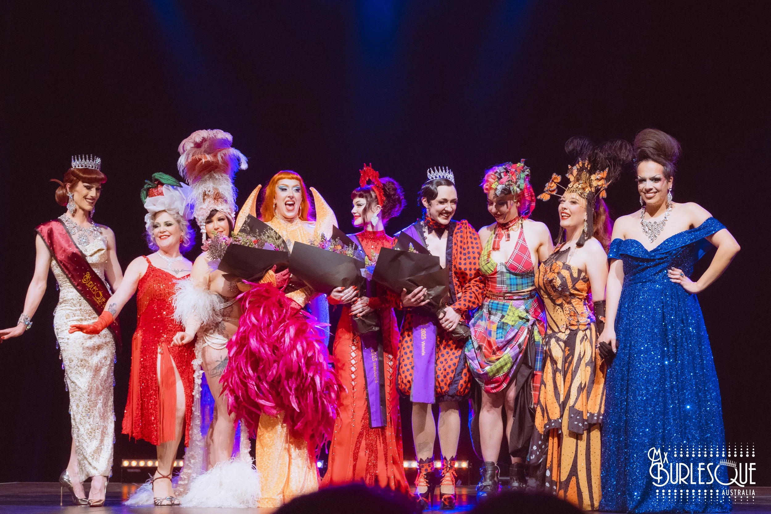A group of women in colorful and extravagant outfits on stage at a fashion or beauty contest. They are smiling and holding bouquets of flowers, with some wearing crowns and elaborate headpieces.