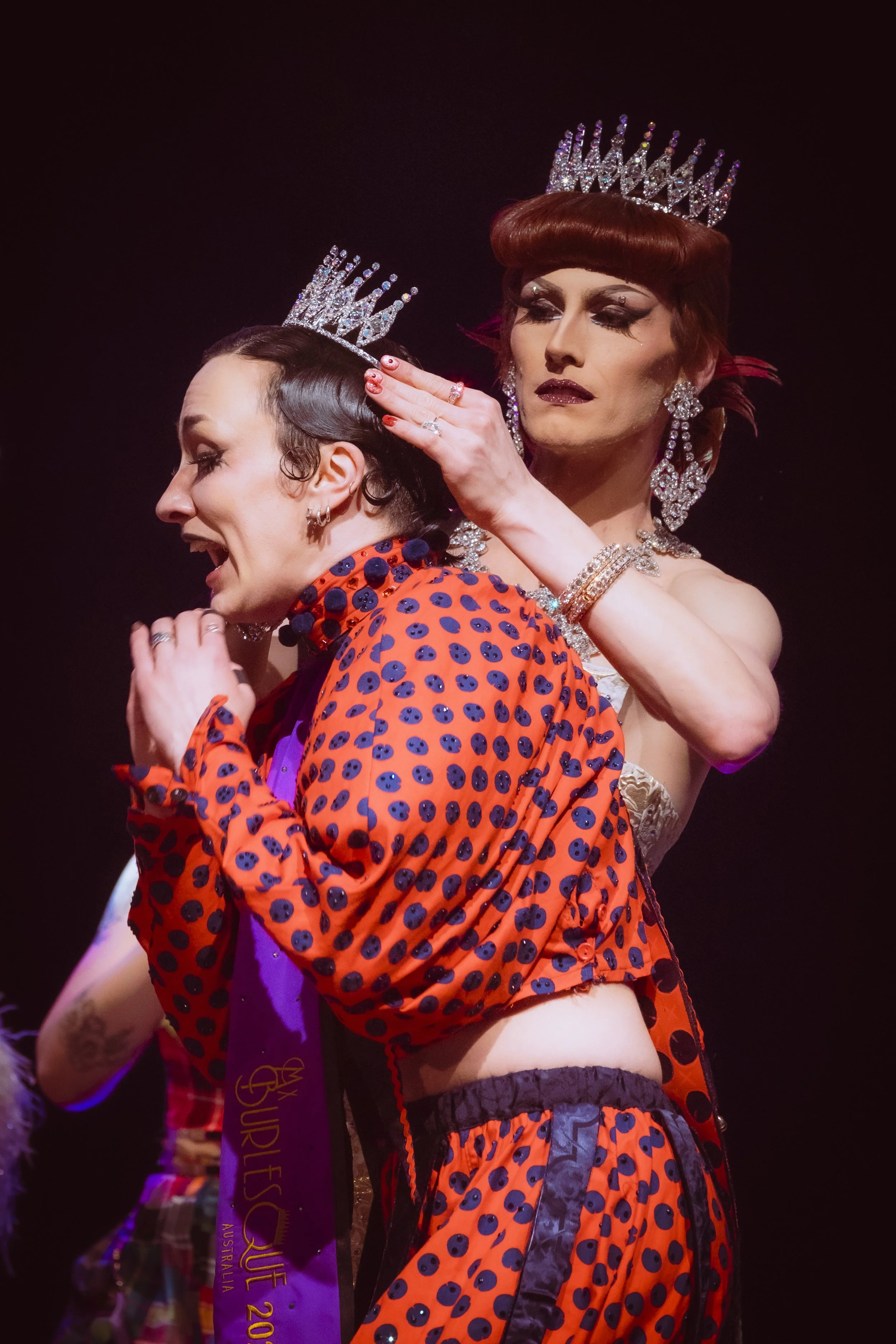 Two burlesque performers one wearing a crown, crown adjustment, vibrant red and black polka dot outfit, in a performance setting.